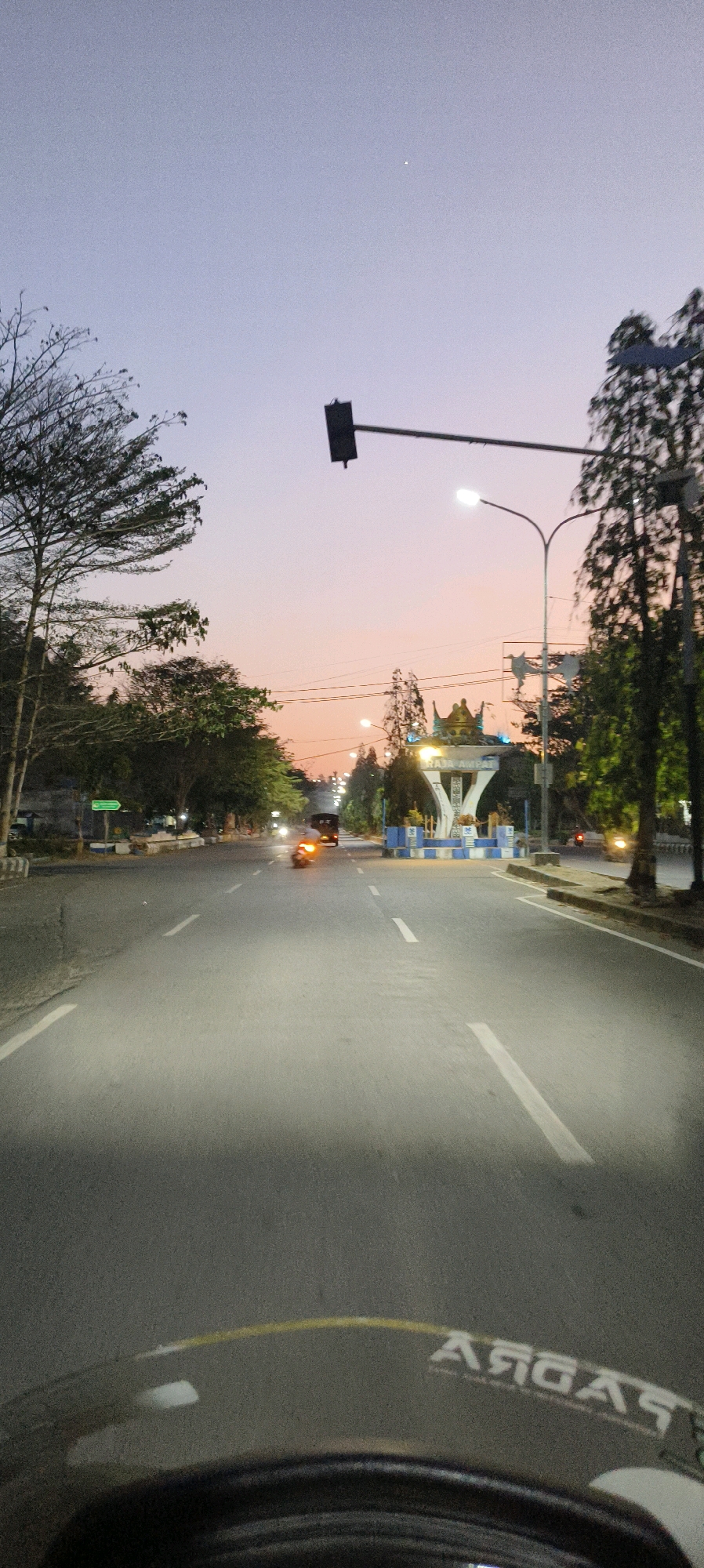 The image depicts an outdoor scene at dusk on a road in Waisai, Indonesia, likely near a landmark. In the foreground, the curved dashboard of a motorcycle is visible, reflecting a yellow stripe and white text that appears to be "RADAG." The road stretches forward, marked with white lane lines, and is illuminated by headlights from approaching vehicles.
On the right side of the road, several trees line the pavement, and a traffic light is positioned above the road. A decorative archway, possibly an entrance to a district or area, is situated in the middle ground, featuring a prominent crown-like sculpture and blue and white checkered barriers. The text "RAJA AMPAT" is visible on the archway.
On the left side of the road, more trees are present, and a green directional sign is partially visible. In the background, the sky displays a gradient of soft purple and pink hues, characteristic of sunset or twilight. A single bright star or planet is visible in the upper left portion of the sky. Traffic on the road includes at least one car and several motorcycles, indicated by their headlights. The overall atmosphere is calm and transitional, marking the end of daylight.
