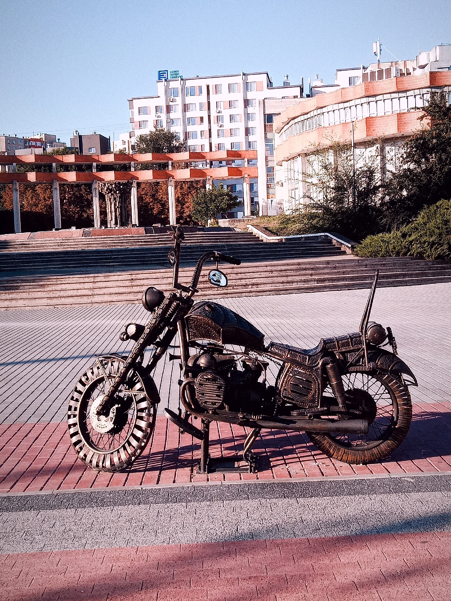 The image depicts a steampunk-style sculpture of a motorcycle on a paved pedestrian area. The motorcycle is made of metal and features intricate details resembling gears and pipes. It is positioned in the foreground, with a blurred background of buildings and a set of stairs leading up to an elevated plaza. The buildings appear to be modern architecture, with some featuring balconies and large windows. The sky is clear and blue. The overall scene suggests an outdoor public space within a city. The location is identified as Chișinău, Moldova, and specifically near the monument to Stephen the Great and Saint.