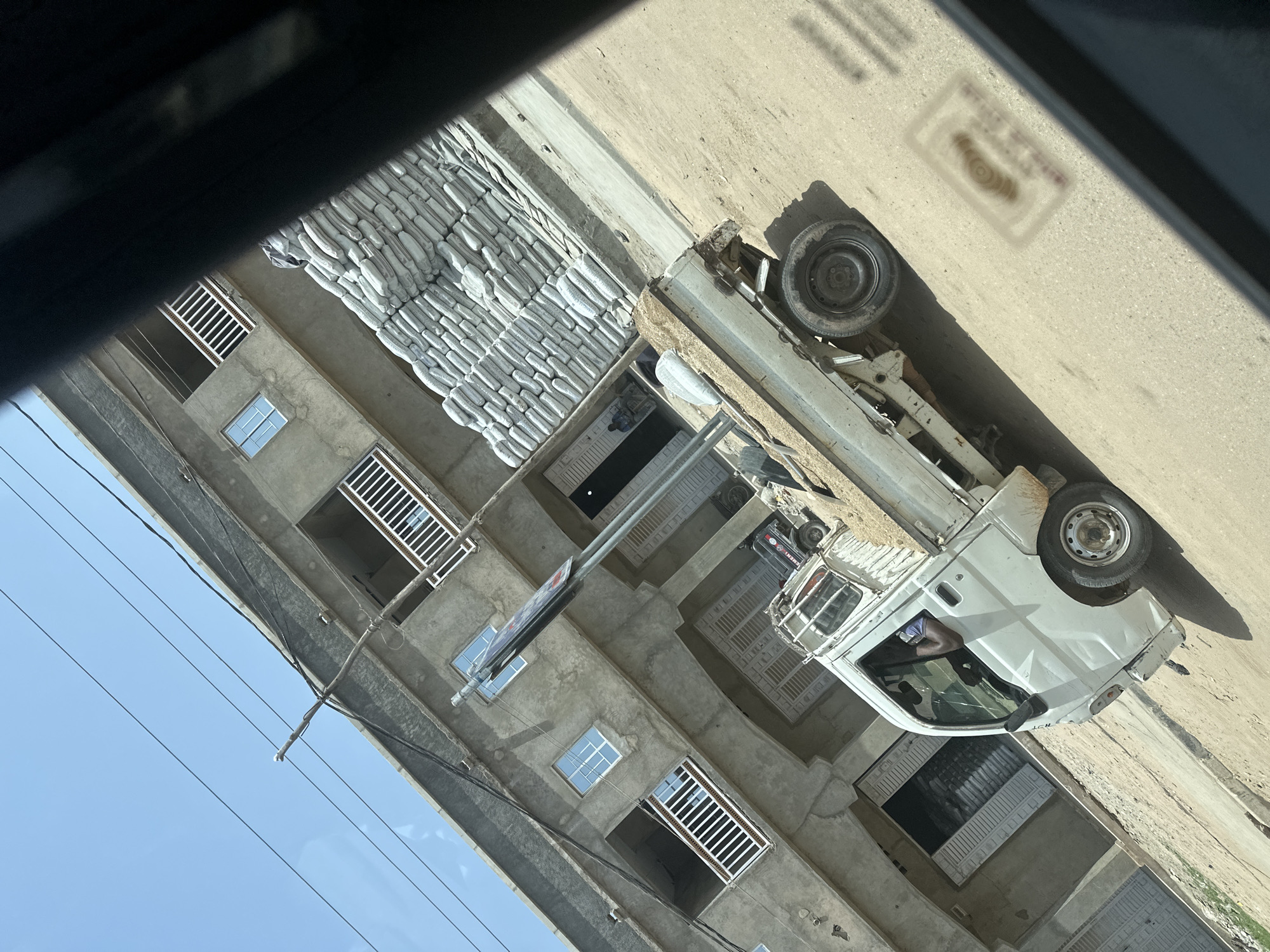 A white pickup truck is parked on a dirt road in front of a multi-story building under construction, its facade made of concrete. The building has several open window frames with white railings and some are fitted with glass windows. A large stack of what appear to be sandbags or cement bags is piled against the side of the building. The pickup truck's bed contains a load of light-colored material, possibly sand or gravel. A man wearing a blue shirt is visible driving the truck. Overhead, power lines are strung across the bright blue sky. The scene is tilted as if captured from a moving vehicle. The general environment suggests a construction or development area within a city.