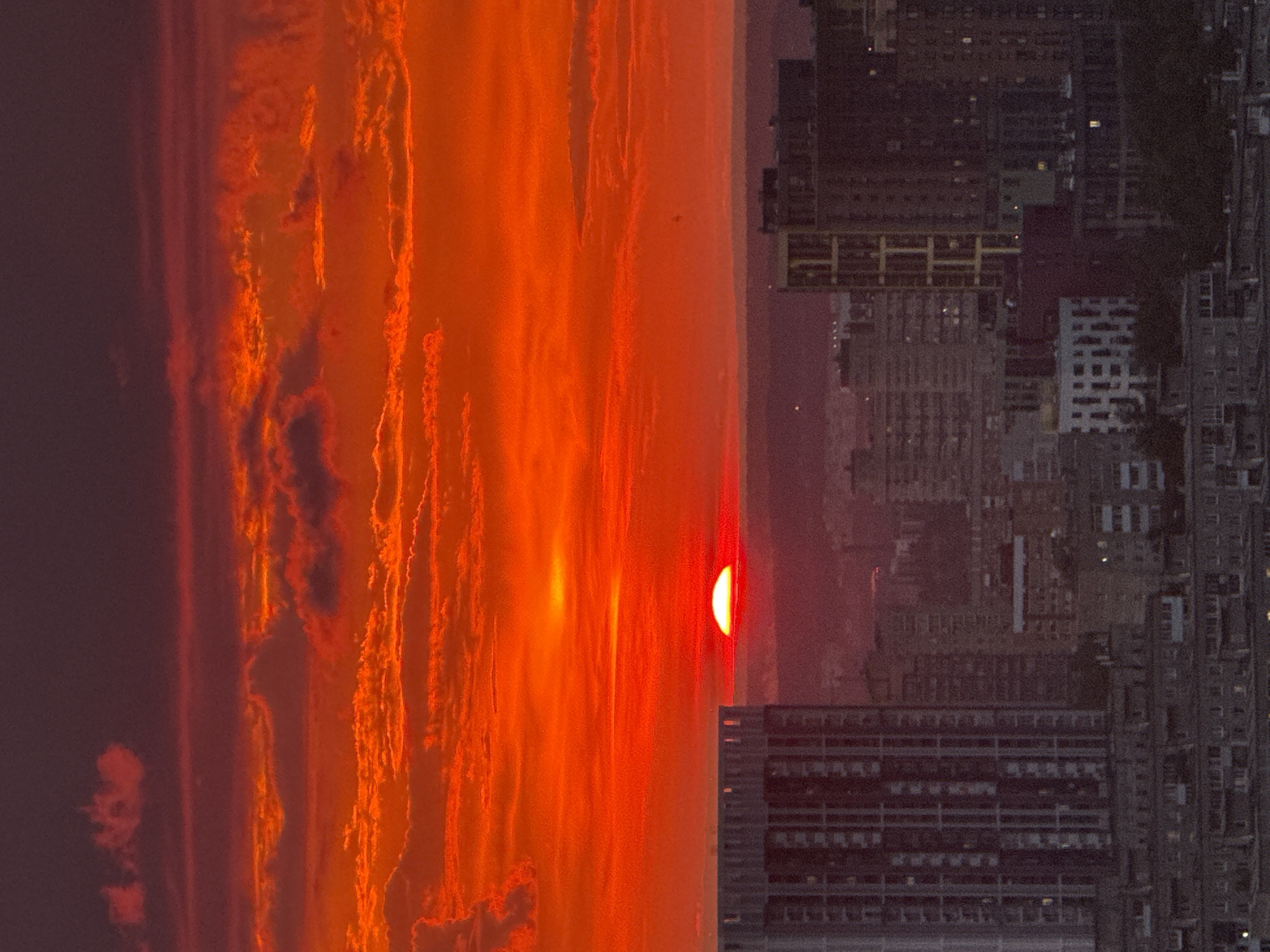 The image shows a vibrant, fiery sunset over the city of Savenki, Russia. The sky is dominated by intense shades of red and orange, with streaks of dark purple at the top. Wispy clouds are illuminated by the setting sun, creating a dramatic and atmospheric effect.
In the foreground, the silhouette of a city skyline is visible. Several buildings, including a tall, modern high-rise on the left and a cluster of shorter, more densely packed buildings on the right, are silhouetted against the bright sky. The details of the buildings are somewhat obscured by the low light and the atmospheric conditions.
The sun itself is a bright, red disc, partially obscured by the horizon, casting a warm glow over the entire scene. The overall impression is one of a powerful natural spectacle occurring above a bustling urban landscape. There are no people or animals visible in the image.