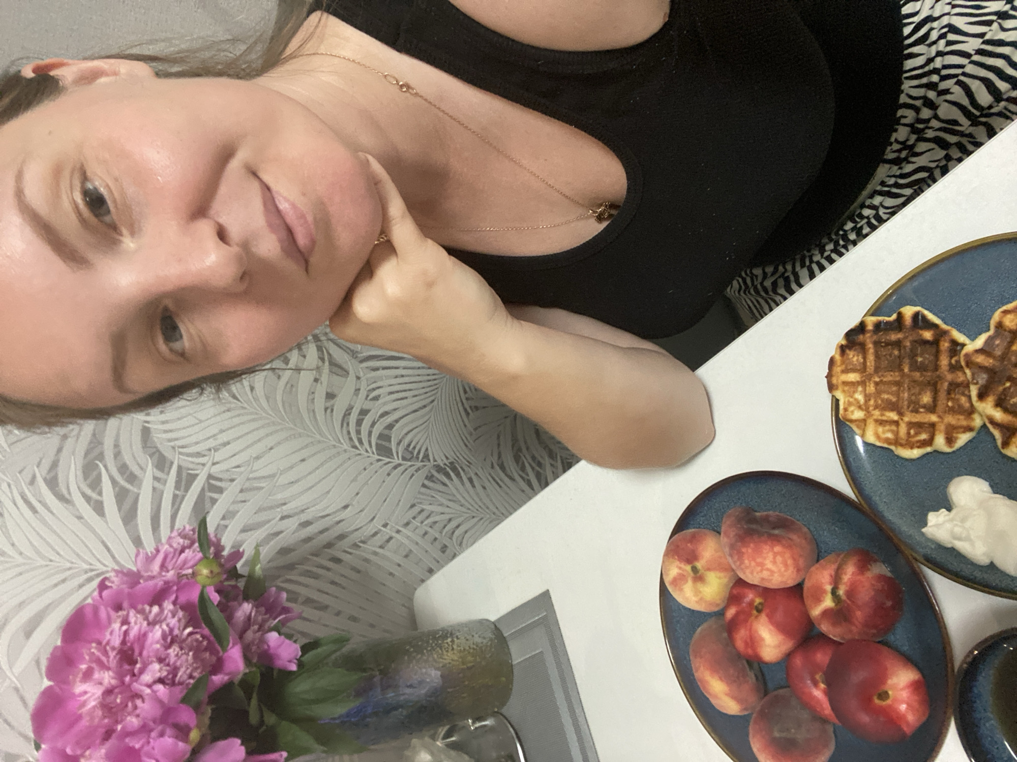 A woman is captured in a close-up selfie at a white table, with a bouquet of pink peonies in a blue vase on her left. In front of her, on a dark blue plate, are several peaches. To her right, another dark blue plate holds two waffles with a dollop of whipped cream. The background features a wall with a white palm leaf pattern wallpaper. The woman is wearing a black tank top and a gold necklace. The scene suggests a meal or snack enjoyed at home. The city of Noviy, Russia is not depicted in this image.