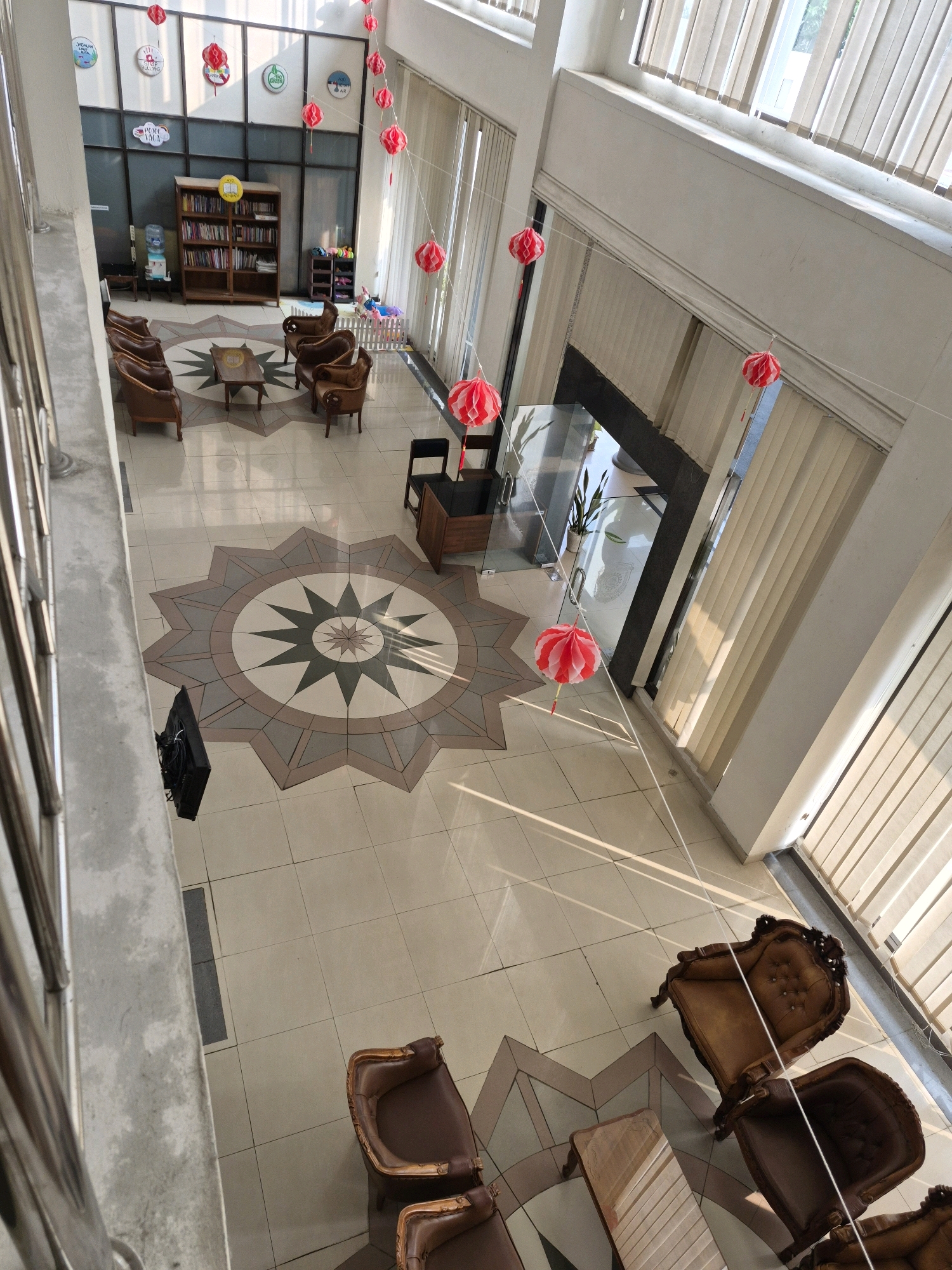 This is a high-angle shot of an interior space, possibly a school or community center, in Purworejo, Indonesia. The scene is dominated by a large tiled floor with a central starburst mosaic design. Red and white paper lanterns are strung across the open atrium, casting shadows.
In the background, a bookshelf filled with books is visible, along with various seating arrangements, including brown leather chairs and a small wooden table. There is also a water cooler and what appears to be a children's play area with a small fence and toys.
On the right side of the image, large windows with vertical blinds are present, allowing natural light to enter the space. In the foreground, a metallic railing and a concrete pillar suggest a walkway or staircase overlooking the main area. A television screen is mounted on the wall in the lower left portion of the image. The overall impression is of a clean, well-lit, and organized indoor environment.