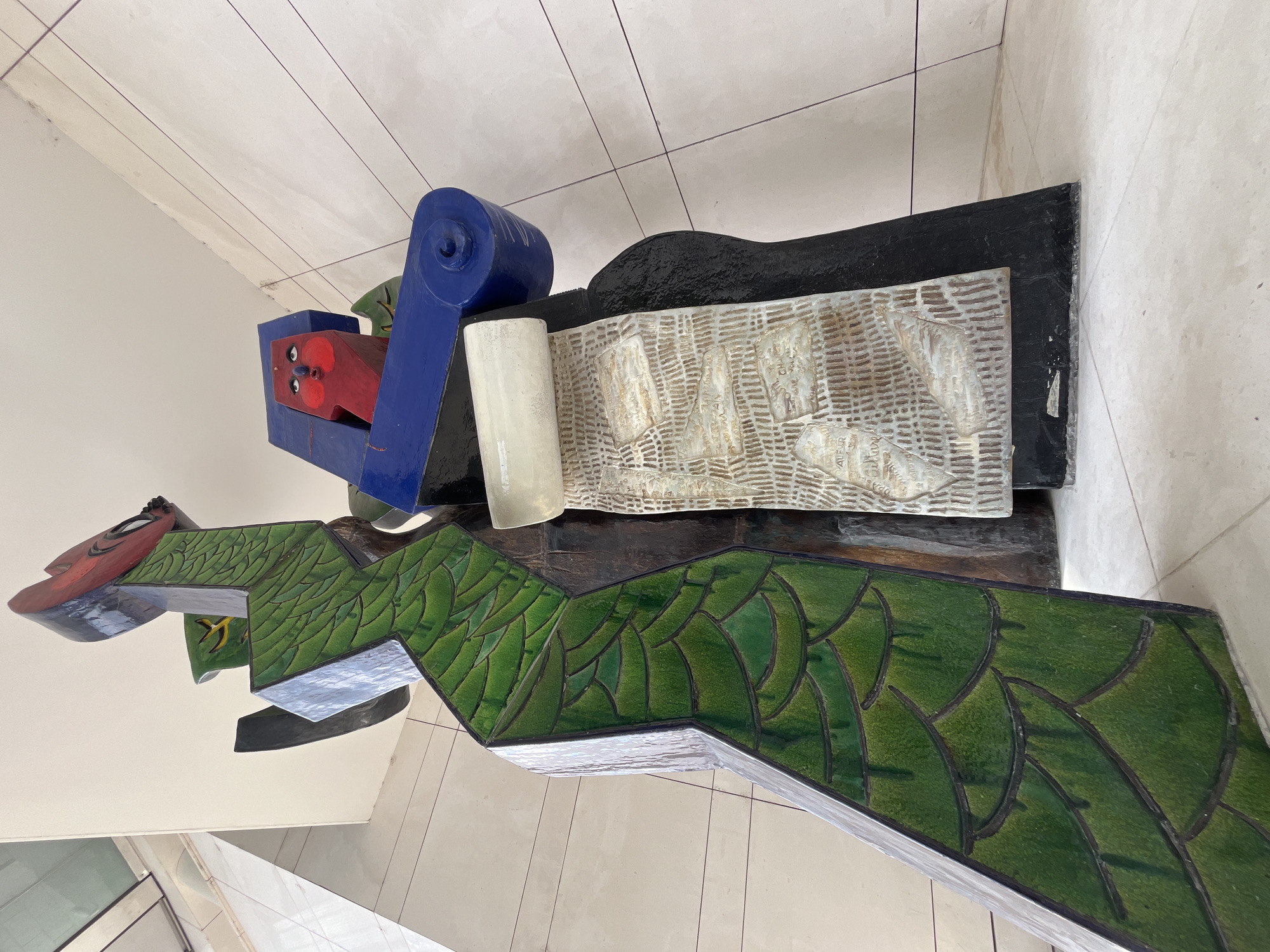 An abstract, multi-colored sculpture is positioned indoors, likely in a public building, indicated by the tiled walls and floor. The sculpture is tall and predominantly features dark brown, green, red, and blue elements. On the left side, a section of the sculpture is covered in a textured, glossy green material arranged in a scale-like pattern. Above this, a red, mask-like face with wide, surprised eyes is visible, set against a blue backdrop. To the right of the face, a cylindrical blue element with a spiral detail protrudes. Suspended from the blue element is a rolled scroll-like object with a textured, off-white surface, displaying what appears to be writing or imagery. The base of the sculpture is dark and textured. The lighting suggests an overhead light source. The environment is clean and modern, with pale beige tiles on the walls and floor.