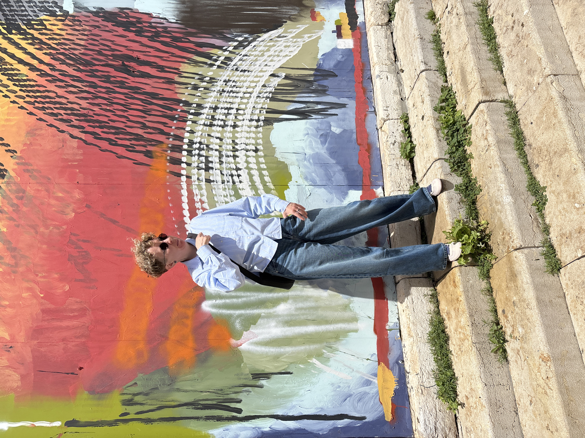 A person with curly hair and sunglasses is lying on their back against a colorful graffiti wall next to concrete steps with some greenery growing between the steps. The person is wearing a light blue striped shirt, blue jeans, and white socks. The location is Tel Aviv, Israel.