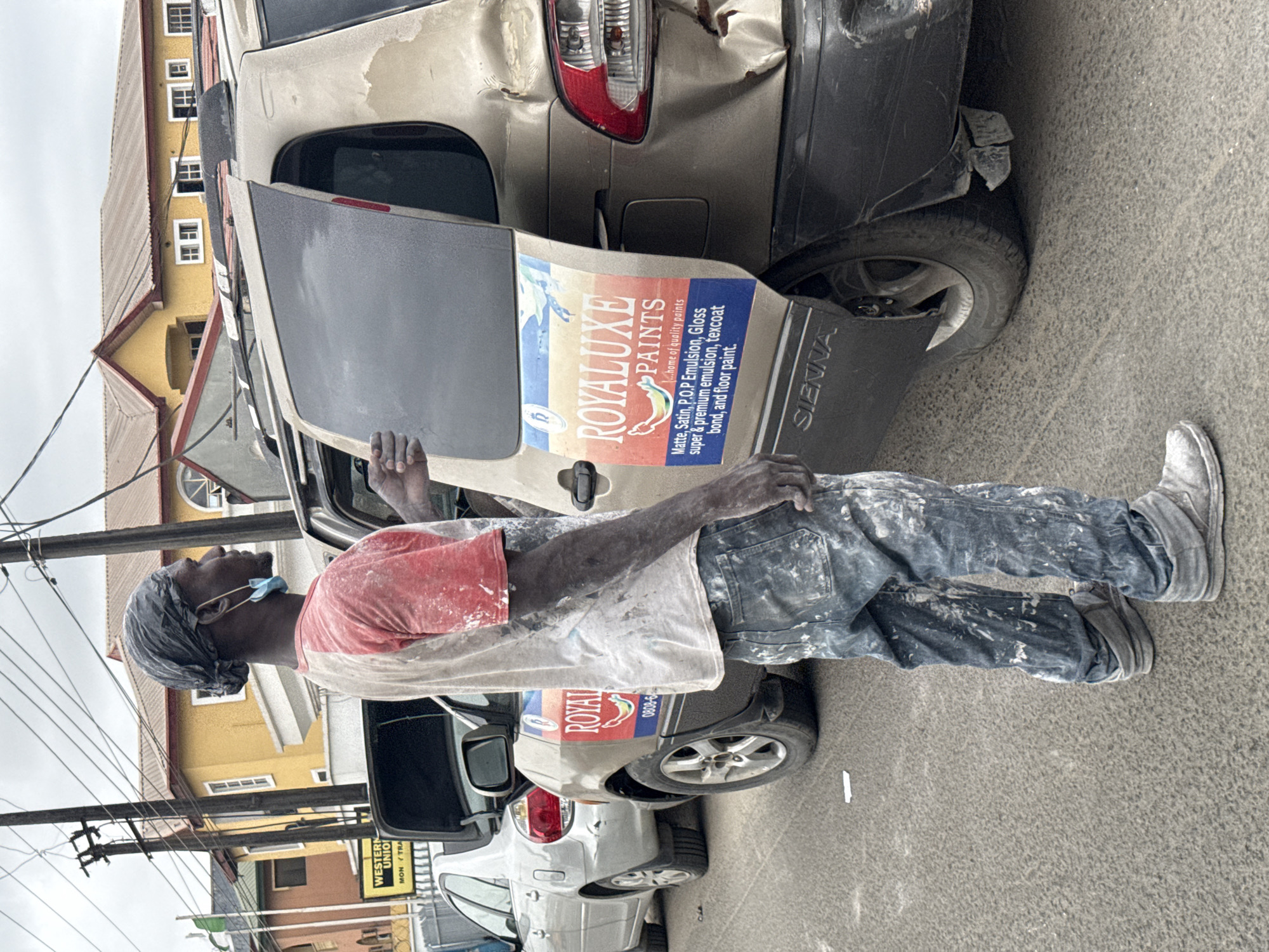 A man, clad in paint-splattered jeans and a red and white t-shirt, stands beside a beige minivan with its rear door ajar. He has a dark cloth wrapped around his head and a light blue face mask hanging from one ear. His body and clothing are covered in white paint splashes, indicative of his profession. The minivan displays a "ROYALUXE PAINTS" advertisement on its rear side panel. In the background, a silver sedan is parked, its trunk open, and further back, buildings with yellow and orange facades are visible. The scene appears to be on a street in Ado, Nigeria.