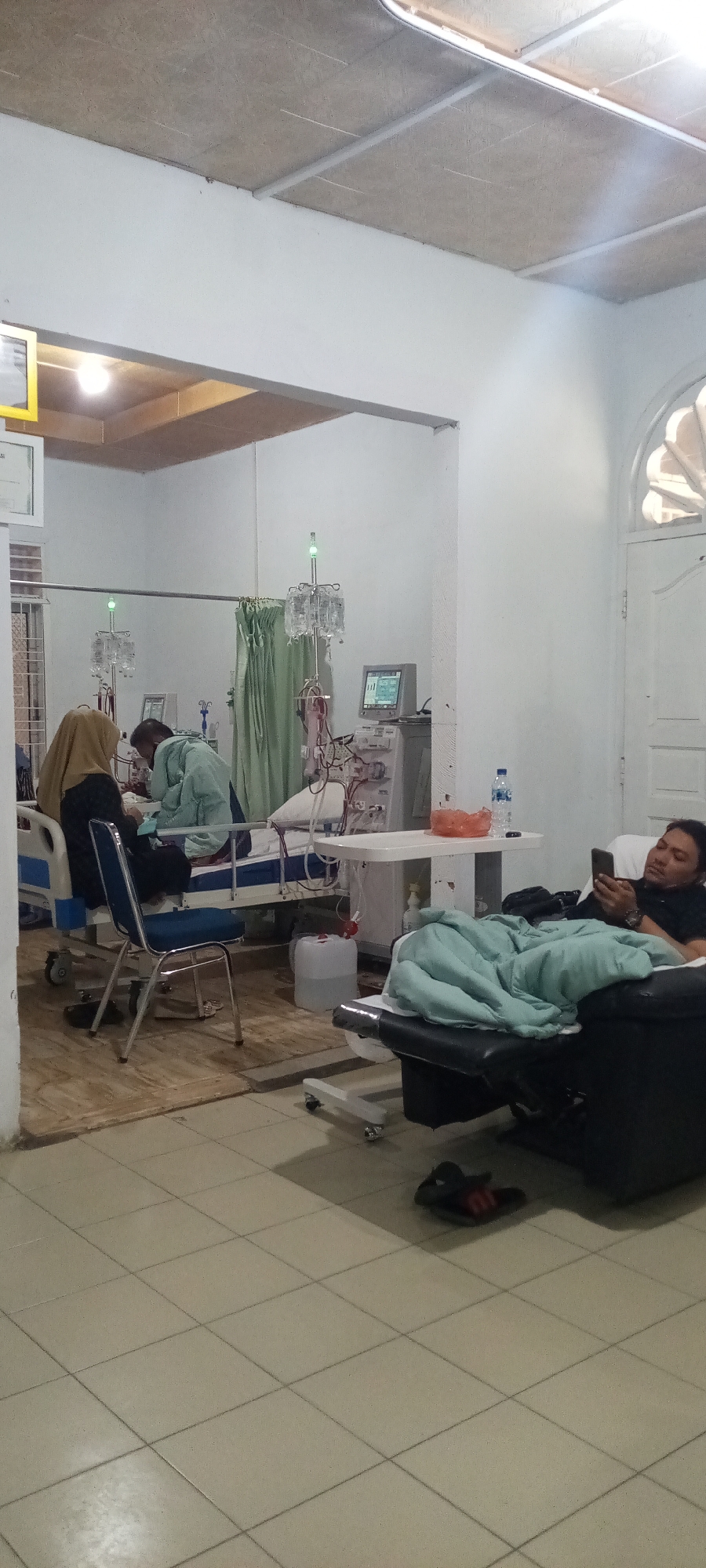 The image depicts a dialysis center in Lhokseumawe, Indonesia. In the foreground on the right, a man is lying on a reclining chair, covered with a light blue blanket, and looking at his smartphone. In the middle ground, two individuals, one wearing a hijab and another in a light green medical gown, are attending to a patient in a hospital bed. Medical equipment, including IV stands with bags of fluid and a dialysis machine with a monitor, are visible. The room has white walls and a tiled floor. A mirrored wall on the left reflects the scene. A door with decorative molding is present on the far right.