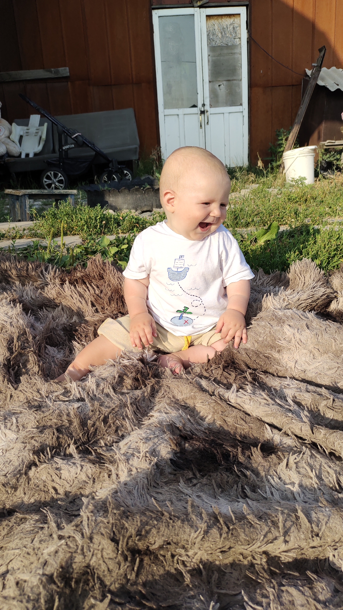 A baby sits on a shaggy, brown blanket outdoors. The baby is bald with light skin and is wearing a white t-shirt with a small sailboat and palm tree graphic and tan shorts. The baby is looking to the right and smiling with their mouth open. The background shows a rustic building with brown siding and a white door. Green grass and some gardening supplies are visible. The lighting suggests it is late afternoon with the sun casting shadows.