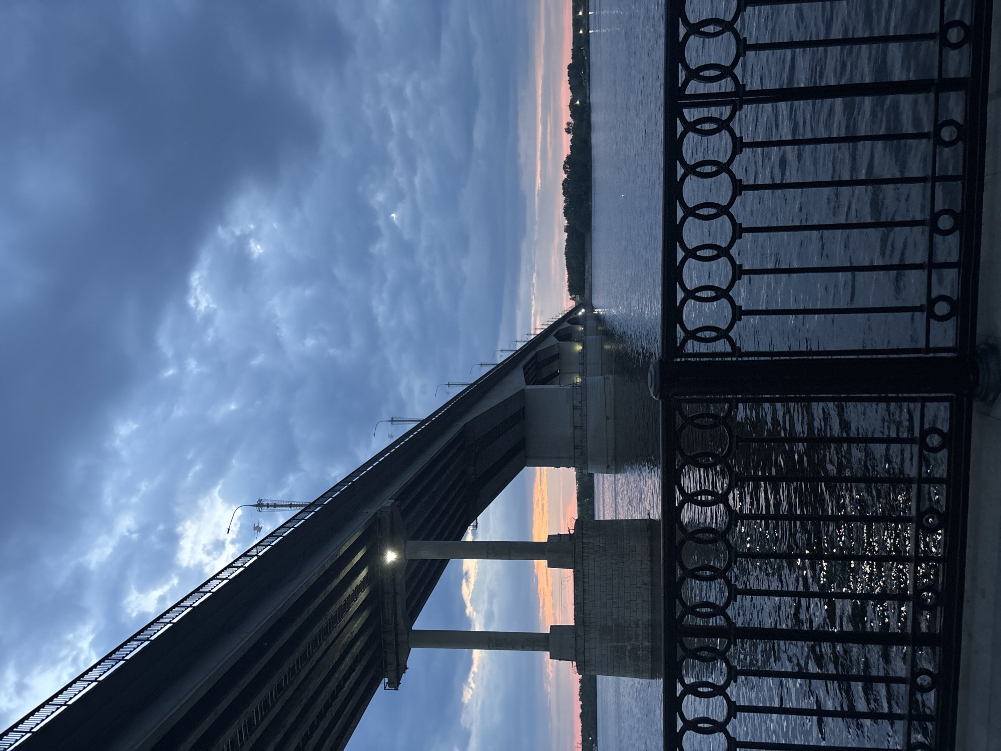 The image shows a view of the Kama Bridge in Perm, Russia, at dusk. The bridge's structure, including supports and lighting, is visible. A decorative metal railing runs along the right side of the frame, partially obscuring the view of the river. The sky is a mix of blues, grays, and pinks from the setting sun. The water reflects the sky and bridge lights. No people are visible.