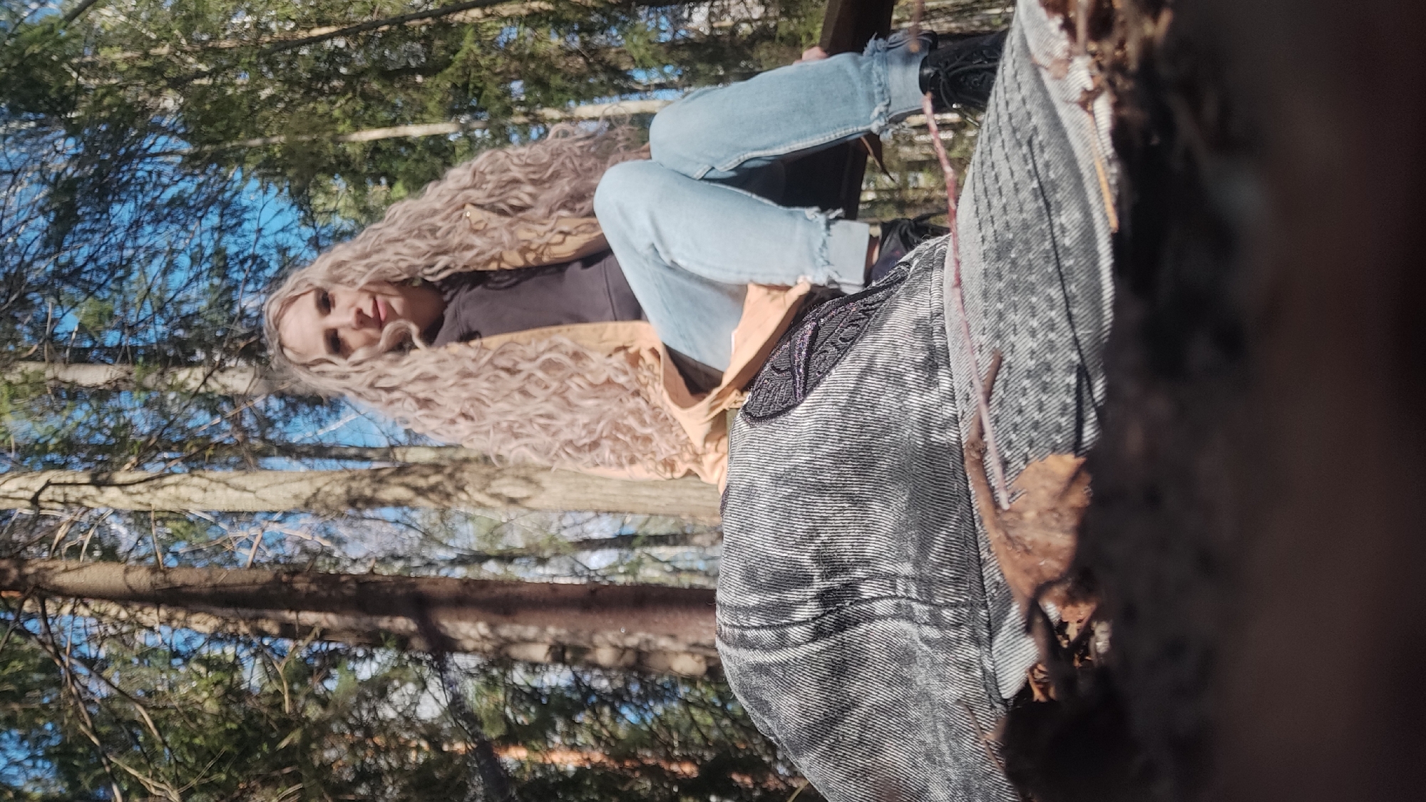 A woman with long blonde, curly hair is reclining, wearing light blue jeans, a dark top, and a tan jacket. She is outdoors, possibly in a wooded area, based on the presence of trees and blue sky visible in the background. The scene includes elements of nature such as trees. Her expression is visible, she is looking towards the camera, possibly smiling.