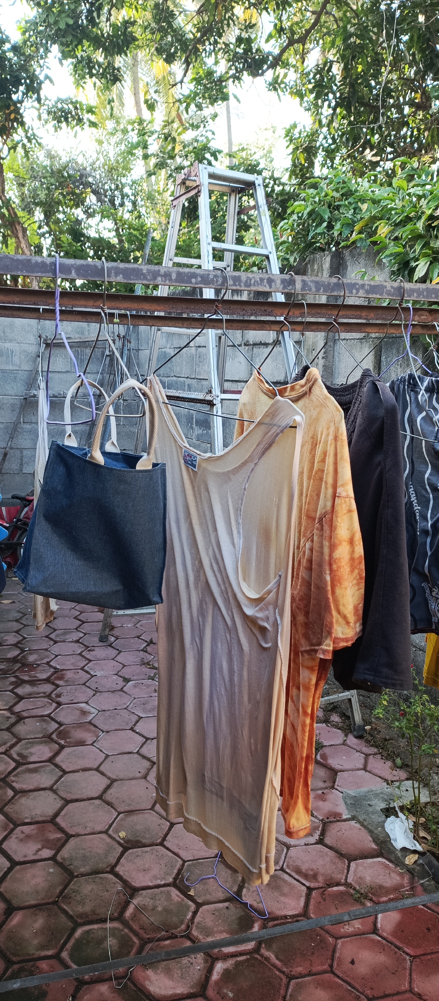 In Arjasa, Indonesia, a metal clothesline hangs in an outdoor area with a brick-tiled floor. Several items of clothing are drying on the line, including a dark blue tote bag with light-colored handles, a sheer, light brown sleeveless top, a short-sleeved shirt with an orange tie-dye pattern, a dark brown garment, and a pair of striped trousers. In the background, a metal ladder stands against a wall, with lush green foliage and trees surrounding the scene. The sky is visible through the trees, suggesting it is daytime.