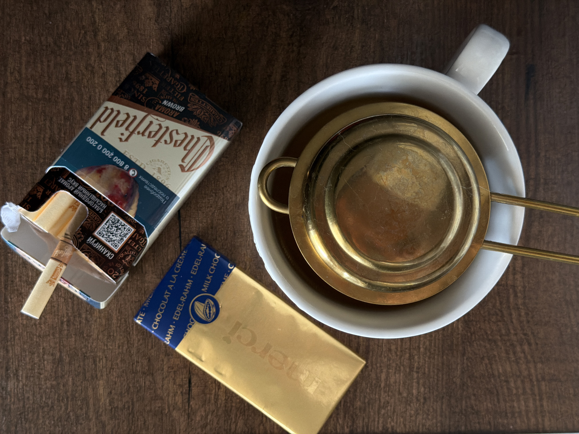 The image shows an open pack of Chesterfield cigarettes, a gold-wrapped Merci chocolate bar, and a white mug with a brass tea strainer inside on a wooden surface. The cigarette pack is partially open, with one cigarette protruding. The chocolate bar is positioned at an angle, with the "merci" logo visible. The white mug contains a tarnished brass tea strainer with two extended handles. The scene appears to be arranged on a dark, possibly varnished, wood surface. No people are visible. The cigarettes are clearly labeled with Russian text indicating the telephone number 8 800 200 0 200 and a warning about the health consequences of smoking. The image was taken in Rossiyskiy, Russia, based on the Russian text on the packaging.
