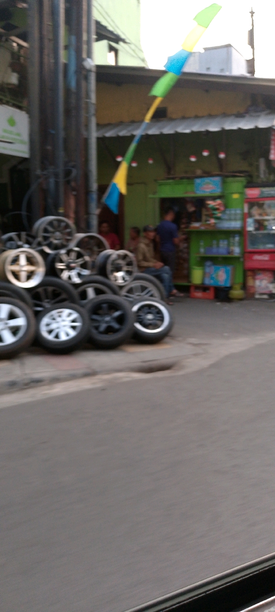 In the city of Bandung, Indonesia, a street-side scene unfolds with a focus on a display of car wheels. The foreground is dominated by a large pile of assorted car wheels, some showing intricate spoke designs and others with simpler patterns. These wheels are stacked haphazardly, with some resting on top of others. Beyond the wheels, a small green kiosk-style shop is visible, with goods displayed on shelves. Several people are present around the shop. One individual, wearing a blue shirt, stands behind the counter of the kiosk, appearing to be engaged in activity within the shop. Another person, in a brown jacket, is seated on the side, facing the shop. Further back, a few more individuals are partially visible, suggesting a bustling commercial area. Above the kiosk, a string of colorful flags or banners, in blue, green, and yellow, is draped, adding a festive element to the scene. The backdrop consists of buildings with pale green and yellow facades, and a gray asphalt road forms the ground. The overall impression is of a busy street corner in a commercial district of Bandung, with a car accessory vendor as a central element.