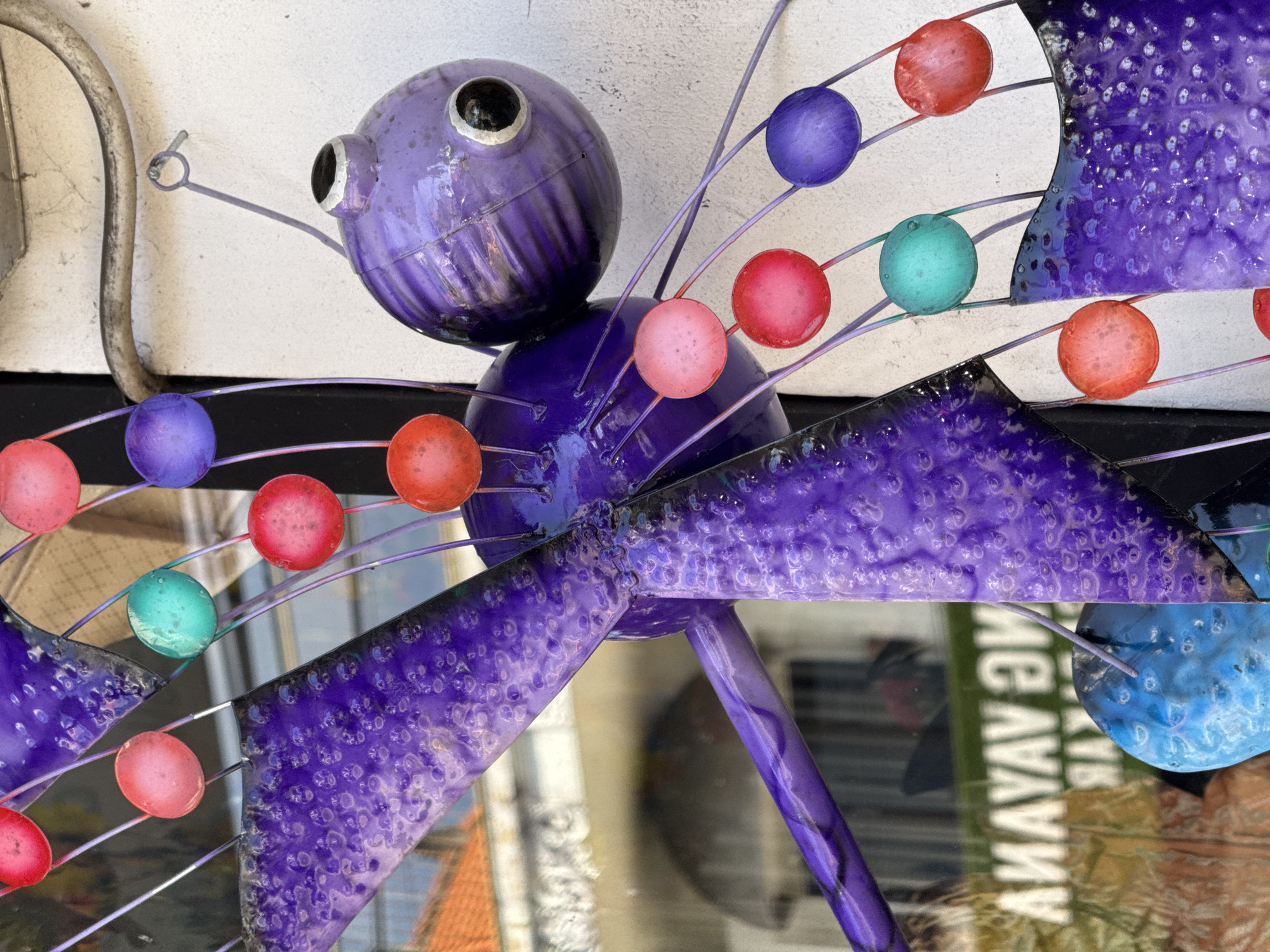 A close-up, low-angle, outdoor shot captures a detailed, metallic purple dragonfly wind spinner. The spinner features large, translucent, bulbous wings with a textured, bubbly surface in shades of purple and blue. Thin, metallic purple wires extend from the dragonfly's body, each suspending a small, round, translucent bead in various colors including red, pink, teal, and purple. The dragonfly's body is segmented and bulbous, with a ribbed texture on the head. The background is a white, textured wall and a glass window reflecting buildings with terracotta roofs. The glass also shows a reversed, white text that reads "AMAYAV EM". The scene is located in Ubud District, Indonesia.