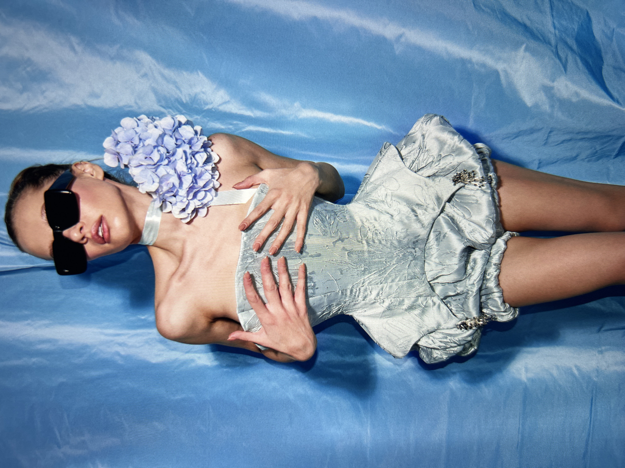 A young woman stands against a blue, draped fabric backdrop. She wears a light blue, strapless corset and ruffled shorts, embellished with a cluster of light purple flowers on her left shoulder and a white ribbon around her neck. She sports oversized, black sunglasses and has her hands clasped over her chest. Her hair is pulled back, and her lips are slightly parted.