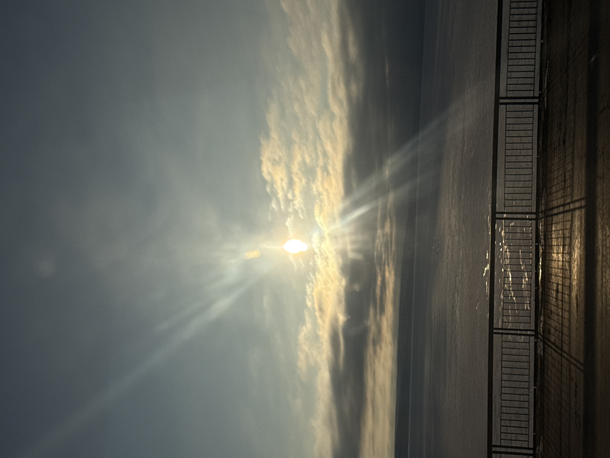 The image depicts a body of water, likely a river or lake, under a sky dominated by clouds and the sun. The sun, positioned slightly off-center, emits bright rays. The water surface reflects the light. A metal railing runs along the right edge of the image, casting a shadow onto what appears to be a walkway or pavement. The location is identified as Пальники, Russia.