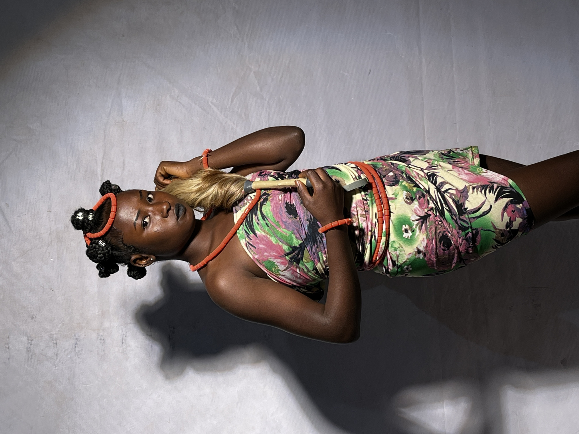 A full-body portrait of a young Black woman, standing and looking towards the viewer against a textured gray backdrop. She has dark skin and her hair is styled in several buns with silver accents, adorned with a red beaded headband. She wears a strapless dress with a vibrant floral pattern in pink, green, and yellow. Red beaded necklaces and bracelets complete her accessories. Her right hand is raised, touching her hair, while her left hand holds a light brown object with a gray handle. A dark shadow of the woman is cast on the left side of the backdrop. The setting is likely a studio or indoor location, given the controlled lighting and backdrop. There are no discernible elements that would specifically indicate the city of Jos, Nigeria.