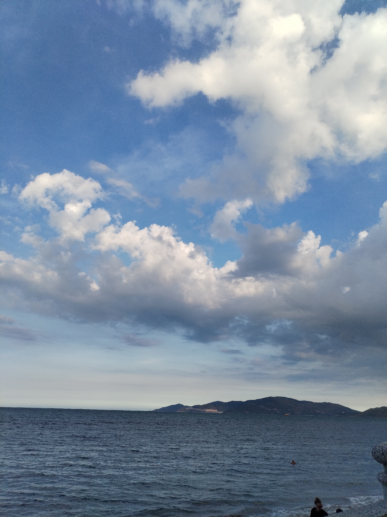 The image depicts a seascape scene with a view of the coastline. In the foreground, there is a beach with waves lapping the shore and a person sitting on the ground. The water stretches out to the horizon, with a distant landmass or island visible in the middle ground. Above, the sky is predominantly blue, scattered with white and gray clouds. The scene appears to be a daytime shot, possibly at the city of Bac Nha Trang Ward, Vietnam.