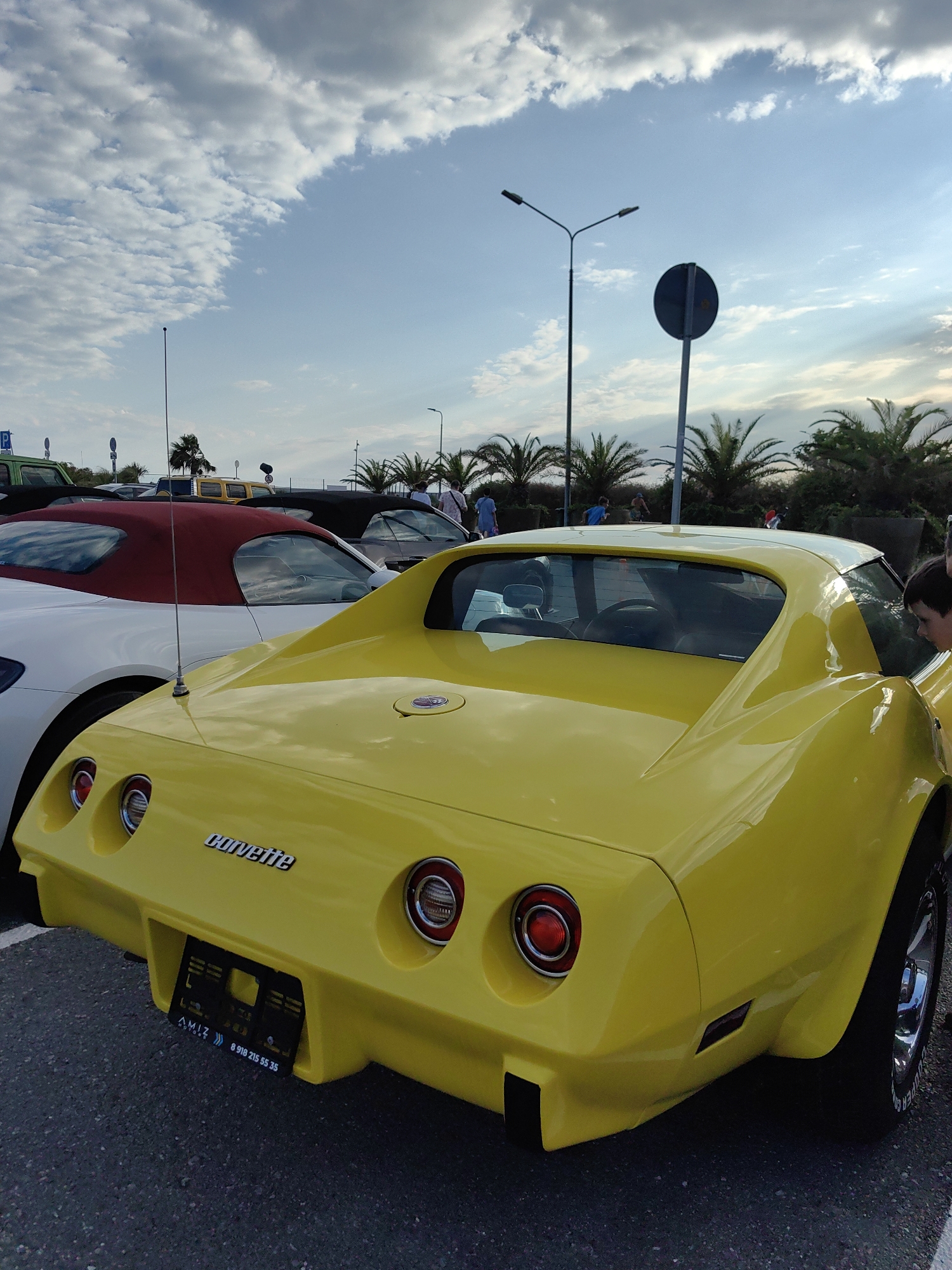 A yellow Corvette is parked in a lot, with a white car with a red top parked to its left. In the background, palm trees line a sidewalk where several people are walking. The sky is cloudy with patches of blue. A sign on the back of the Corvette reads "Corvette." A license plate is visible below the Corvette's taillights with text that appears to be "AMIZ" and a phone number. The scene is outdoors, suggesting it could be a parking lot or a street. The location is identified as Sirius, Russia.
