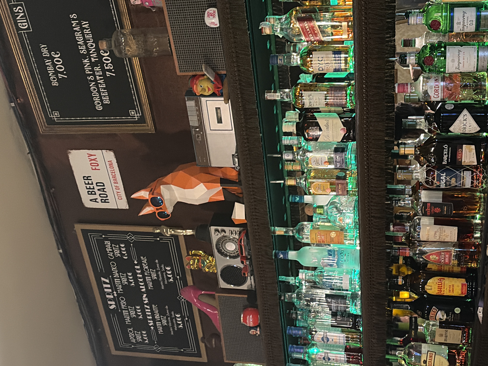 The image depicts a bar interior with a focus on a shelf filled with various bottles of alcohol. The bar has a vintage aesthetic, with chalkboards displaying drink menus and decorative items. One chalkboard lists "SPRITZ" with different variations like "APEROL," "MARTINI FIERO," "MARTINI BIANCO," and "CAMPARI," all priced at 5.00€. Another section of the chalkboard is dedicated to "SPRITZ SIN ALCOHOL," featuring "MARTINI VIBRANTE" and "MARTINI FLOREALE," also priced at 5.00€. A separate framed chalkboard to the right is titled "GINS" and lists "BOMBAY DRY" at 7.00€ and "GORDON'S PINK, SEAGRAM'S BEEFEATER, TANQUERAY" at 7.50€.
Above the bar shelf, several decorative objects are visible, including a pink high-heeled shoe, a golden lucky cat figurine, a vintage-style radio, and a stylized paper-craft fox wearing sunglasses. A sign reading "A BEER ROAD FOXY CITY OF BARCELONA" is mounted on the wall.
The main focus of the image is the illuminated bar shelf below, which is stocked with an extensive collection of liquor bottles. These bottles are lit from below with green and blue lights, creating a vibrant atmosphere. Brands like Martini, Jägermeister, Grey Goose, Dewar's, Johnnie Walker Red Label, Tanqueray, Gordon's, and Hendrick's are identifiable among the many bottles. The shelves are organized with multiple rows of bottles, suggesting a well-stocked bar. The background is a dark, textured wall, contributing to the moody ambiance of the establishment. The image appears to be taken from a slightly elevated perspective, looking down at the bar.