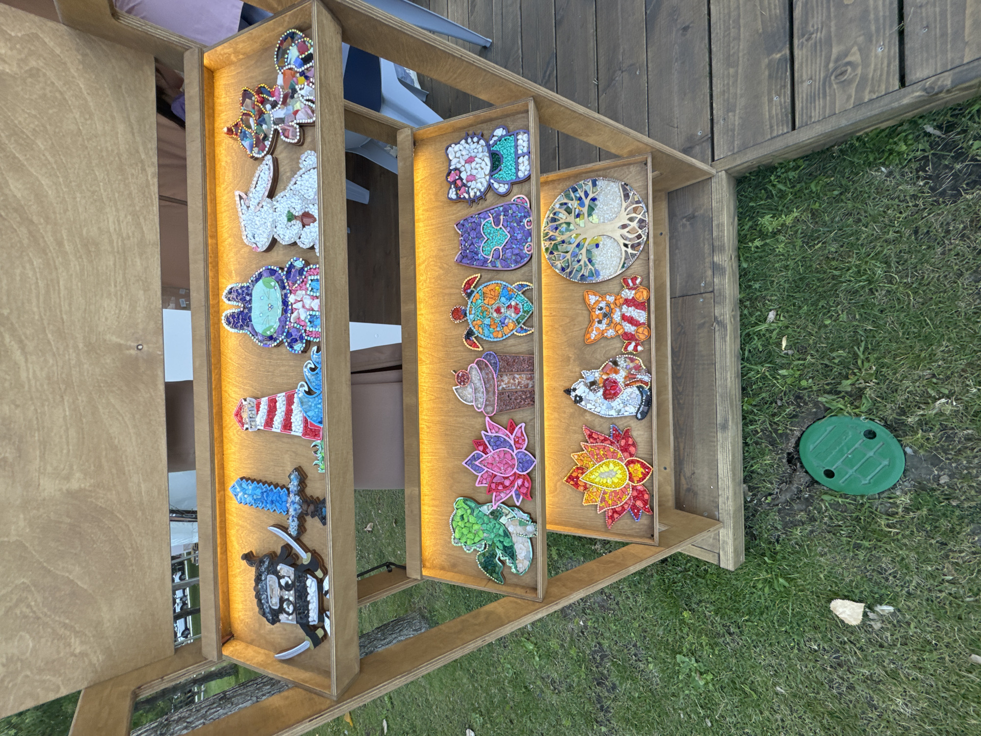The image depicts a wooden display shelf filled with colorful, mosaic-style decorative items. The shelf is outdoors, with grass and wooden planks visible in the foreground, suggesting a park or outdoor market setting. The items on the shelves are diverse, including a ninja figure, a sword, a lighthouse, rabbits, a frog, a cupcake, a turtle, cats, a lotus flower, a fox, and a tree of life. The items appear to be handcrafted, possibly made from beads or small stones, and are arranged in rows on the lit shelves. There are no people clearly visible in the image, but a reflection in the top shelf suggests the presence of people in the background. The overall scene suggests a display of artisanal crafts for sale or exhibition. There is no specific indication of the location being in Anapa, Russia.