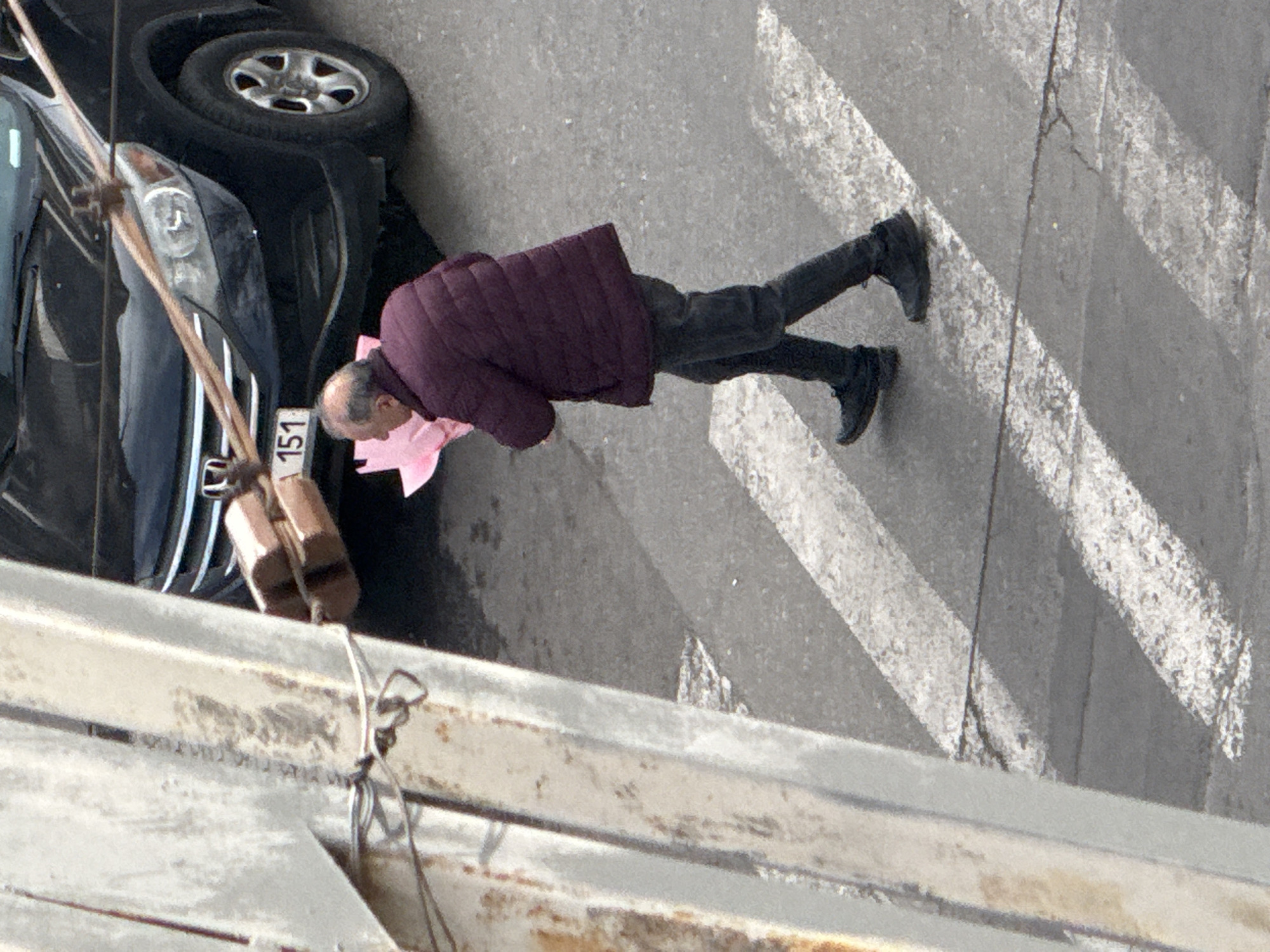 An elevated view captures an urban street scene in Yerevan, Armenia. An adult male, observed from the upper torso down, walks from left to right across a pedestrian crosswalk marked with white diagonal stripes on asphalt. He wears a dark maroon puffer jacket, dark trousers, and dark athletic shoes, and carries a light pink wrapped item, consistent with a floral bouquet or gift, clutched in his left arm against his chest. In the background, a black Honda CR-V SUV is parked or stationary, with its front bumper, left headlight, and a partial view of its front tire visible. The vehicle's license plate displays "151" with Armenian country identification. A large, light-colored metal structural column, exhibiting signs of wear and rust, occupies the left foreground, partially obscuring the scene. A dark cable or wire extends diagonally across the frame, suspended by two small, brown cylindrical insulators or spacers, passing over the SUV's front.