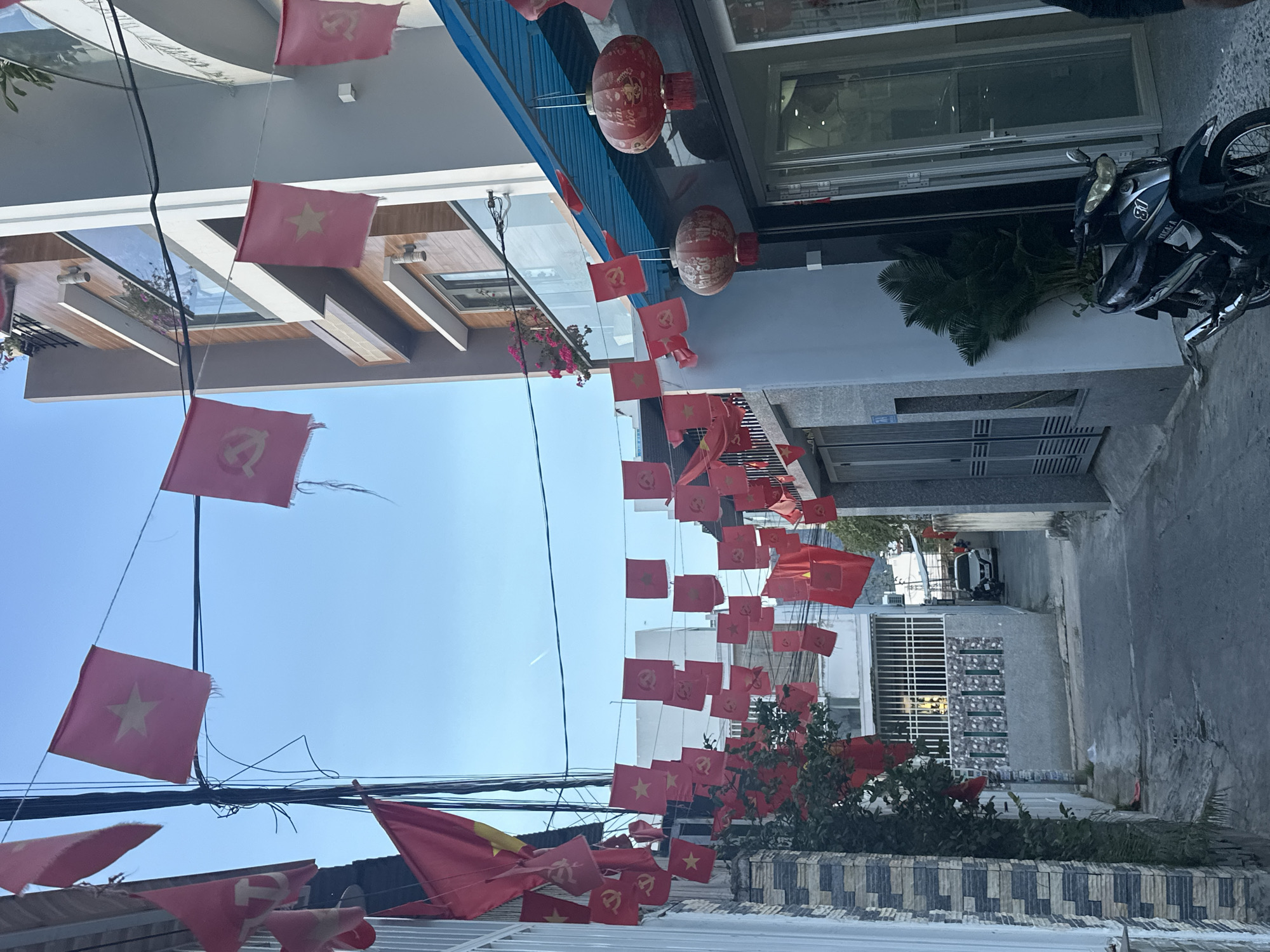 A street in Bac Nha Trang Ward, Vietnam is decorated with strings of red flags, some with a gold star (national flag of Vietnam) and some with a hammer and sickle (communist party flag). Two red lanterns hang above a building with a blue awning. A motorcycle is parked outside a building to the right. The sky is blue.