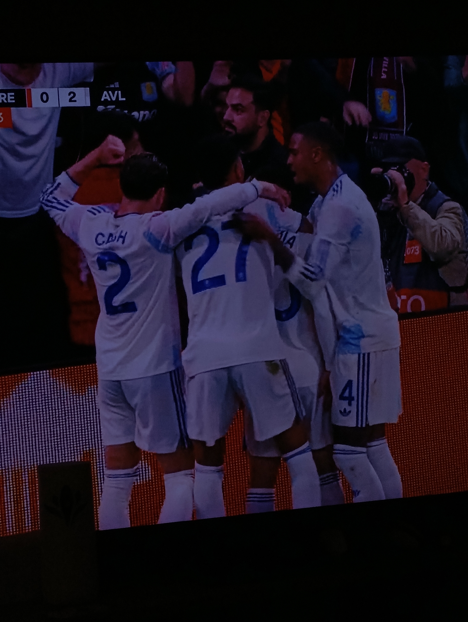 The image shows a soccer match, with the score showing "0-2" in favor of AVL (Aston Villa). Four players in white jerseys with blue numbers are celebrating a goal. The numbers on their backs are 2, 27, and 4 (partially visible). There are other people in the background, including a person with a camera. A sign with "VILLA" is also visible. The game is taking place on a soccer field. The location and city of Plovdiv, Bulgaria are not evident in the image.