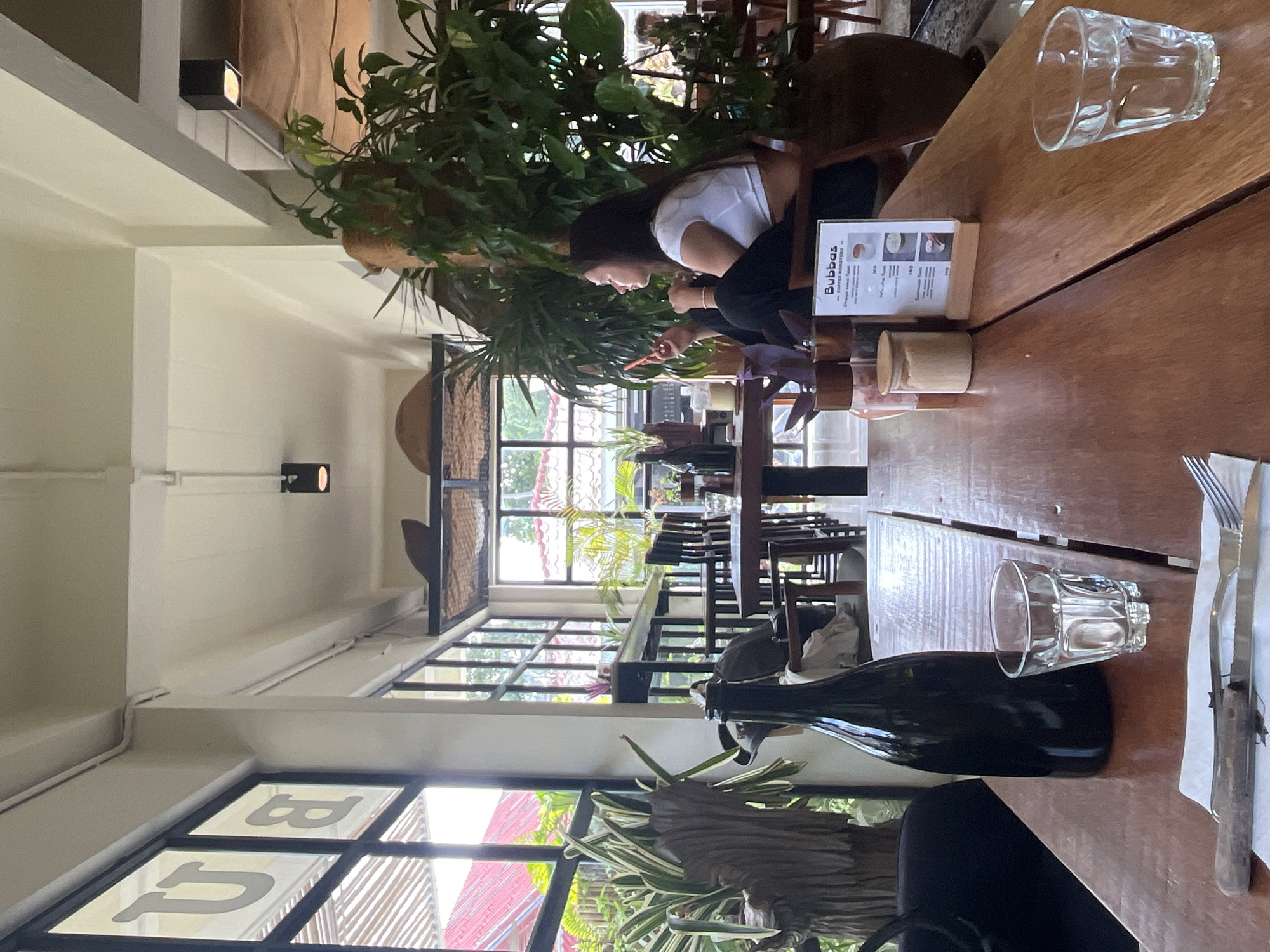 This image depicts the interior of a cafe in Koh Phangan, Thailand, with a focus on a wooden table in the foreground. On the table, a tall, dark bottle and two glasses of water are visible. To the right, a menu stand with the text "Bubbas COFFEE ROASTERS" is placed. A fork and knife are laid out on a napkin. In the background, a young woman with long dark hair, wearing a white crop top and black pants, sits at a table, looking at her phone. She is positioned near large potted plants. The cafe has large windows with black frames that allow natural light to enter, illuminating the dining area. Through the windows, glimpses of outdoor greenery and a red-tiled roof can be seen. The cafe's ambiance suggests a relaxed, modern setting with a botanical theme, indicated by the abundant plant life. The overall scene captures a moment of leisure within a cafe environment.