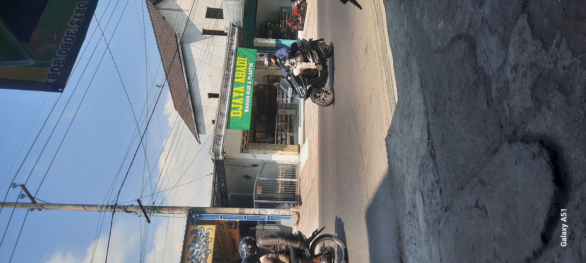 A street scene in Selopuro, Indonesia shows a street with several motorcycles. A person wearing a helmet and a dark jacket is riding a motorcycle on the left side of the image. On the right side, another person wearing a helmet and a light-colored jacket is riding a dark-colored scooter. In the background, there are shops and buildings. One shop has a green banner that reads "DJAYA ABADI BAHAN KUE & PLASTIK". Another shop has a sign that reads "UD. RIZQI JAYA MOTOR". There are utility poles and power lines overhead, and the sky is blue with some clouds. The street appears to be in moderate condition with some visible wear and tear.