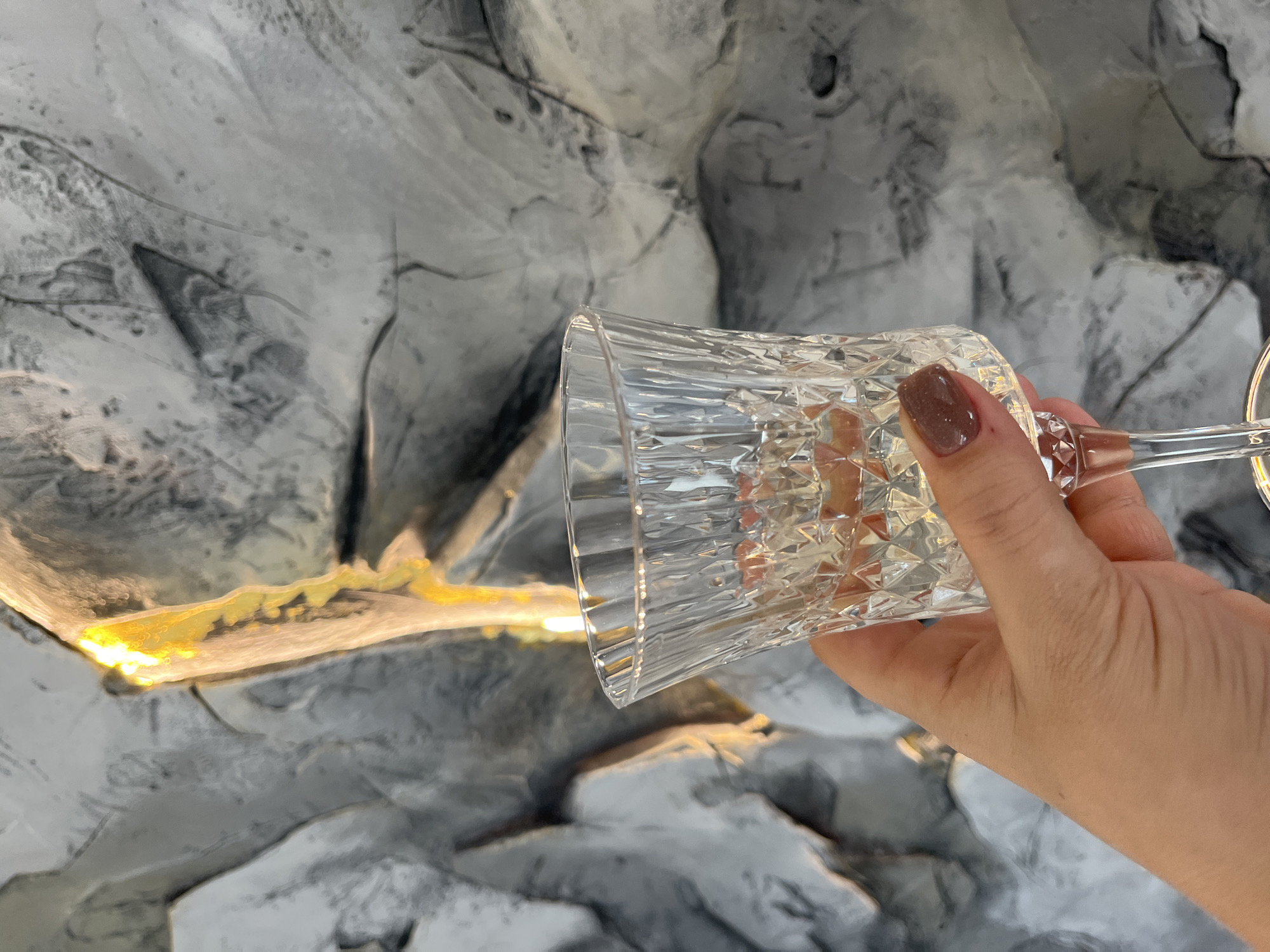 A close-up, high-angle shot depicts a light-skinned hand, likely female, holding a clear, stemmed glass. The hand is positioned in the lower-left quadrant of the frame, with the thumb and fingers gripping the bowl of the glass. The fingernails are short and uniformly coated with a glossy, solid brown nail polish. The glass features a faceted, diamond-cut pattern on its bowl and contains a clear liquid, approximately halfway full. The background consists of a highly textured wall or surface rendered in shades of gray and white, with prominent irregular ridges and valleys, suggesting a decorative plaster finish or a natural stone facade. A vertical band of bright yellow-orange light emanates from the upper-middle section of the background, illuminating parts of the textured surface and creating a strong contrast with the predominantly cool-toned environment. The scene is located within a building in Новая Чернушка, Russia.
