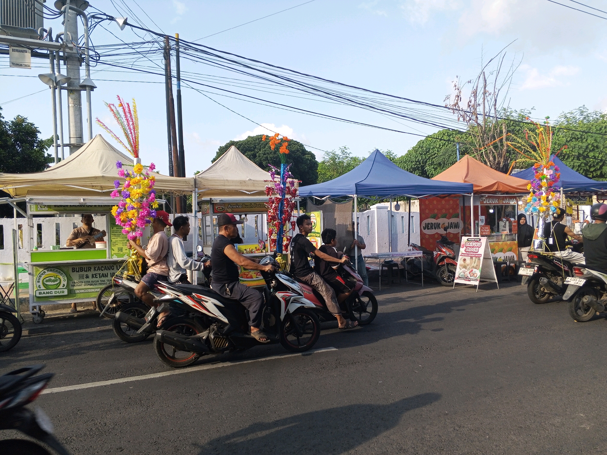 In Tamansari, Indonesia, a street scene unfolds under a bright sky with scattered clouds, likely during the daytime. The foreground is dominated by an asphalt road, with shadows cast by the sun indicating a late afternoon or early morning setting. Several motorcycles are present, some parked and others in motion.
Along the side of the road, a row of market stalls operates. These stalls are covered by various colored tents, including beige, blue, and orange. The stalls appear to be selling food and drinks, with signage visible for products like "BUBUR KACANG IJO" (mung bean porridge), "ANeka BUAH POTONG" (assorted cut fruits), and "JERUK JAWA" (Javanese oranges). Decorative elements, such as colorful artificial flower arrangements, are displayed, adding a festive touch.
Multiple individuals are engaged in various activities. Some are manning the stalls, serving customers or preparing goods. Others are riding motorcycles, navigating the street. One motorcycle has three people on it, suggesting a shared ride. The people are dressed in casual clothing, typical for the region. In the background, trees and utility poles with numerous overhead wires suggest an urban or semi-urban environment. A large transformer box is also visible on one of the utility poles. The overall atmosphere is that of a bustling local market or street fair.