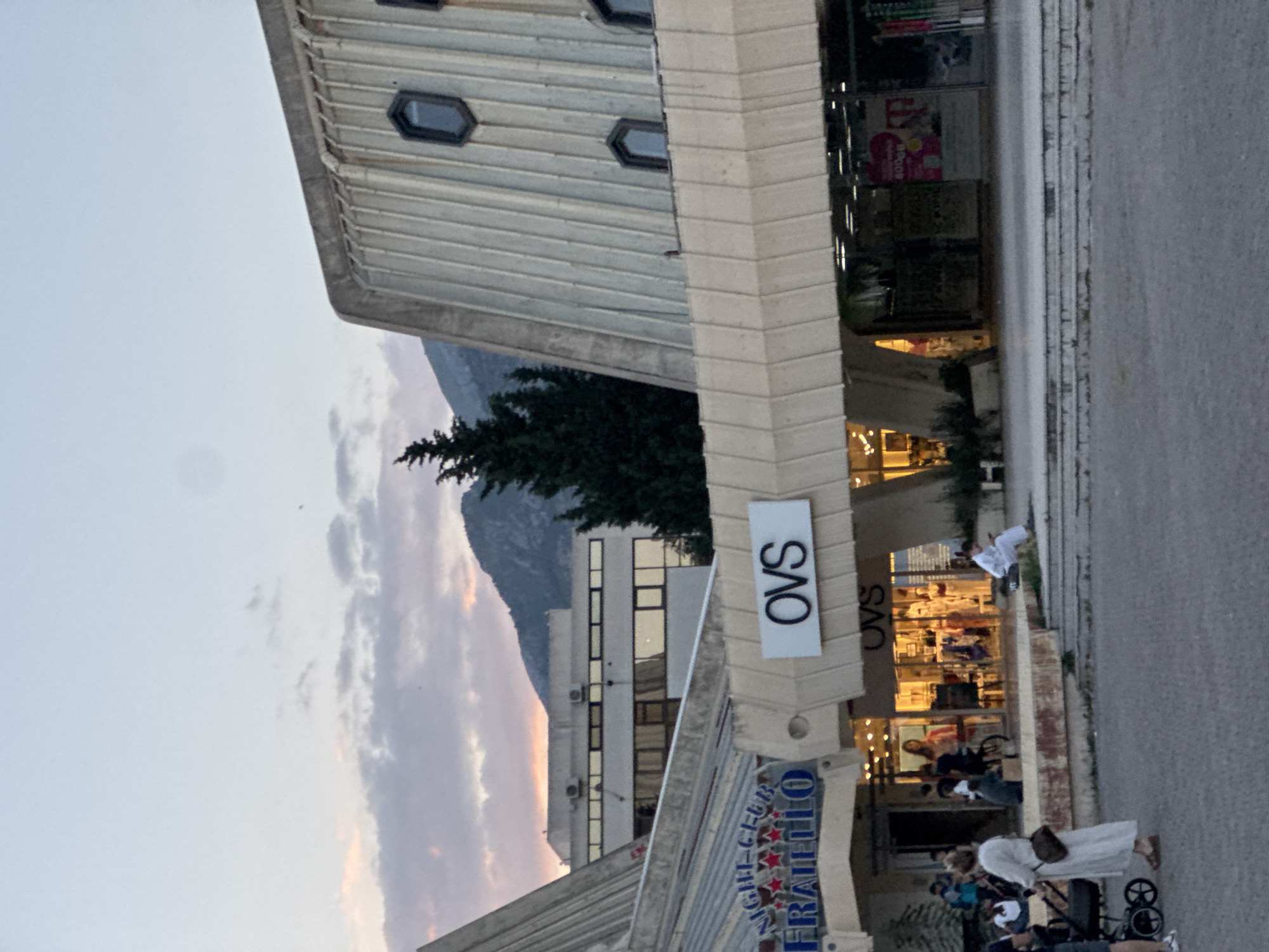 The image captures a street scene in Bar, Montenegro, facing a building with a retail space. An "OVS" sign, visible on a protruding architectural element, indicates one of the businesses. People, including families with strollers, are present near the storefront, suggesting pedestrian activity. In the background, another building can be seen, and further behind that, a mountain is visible against a sky with clouds. The lighting indicates that it is dusk or dawn. The image is taken from street level.