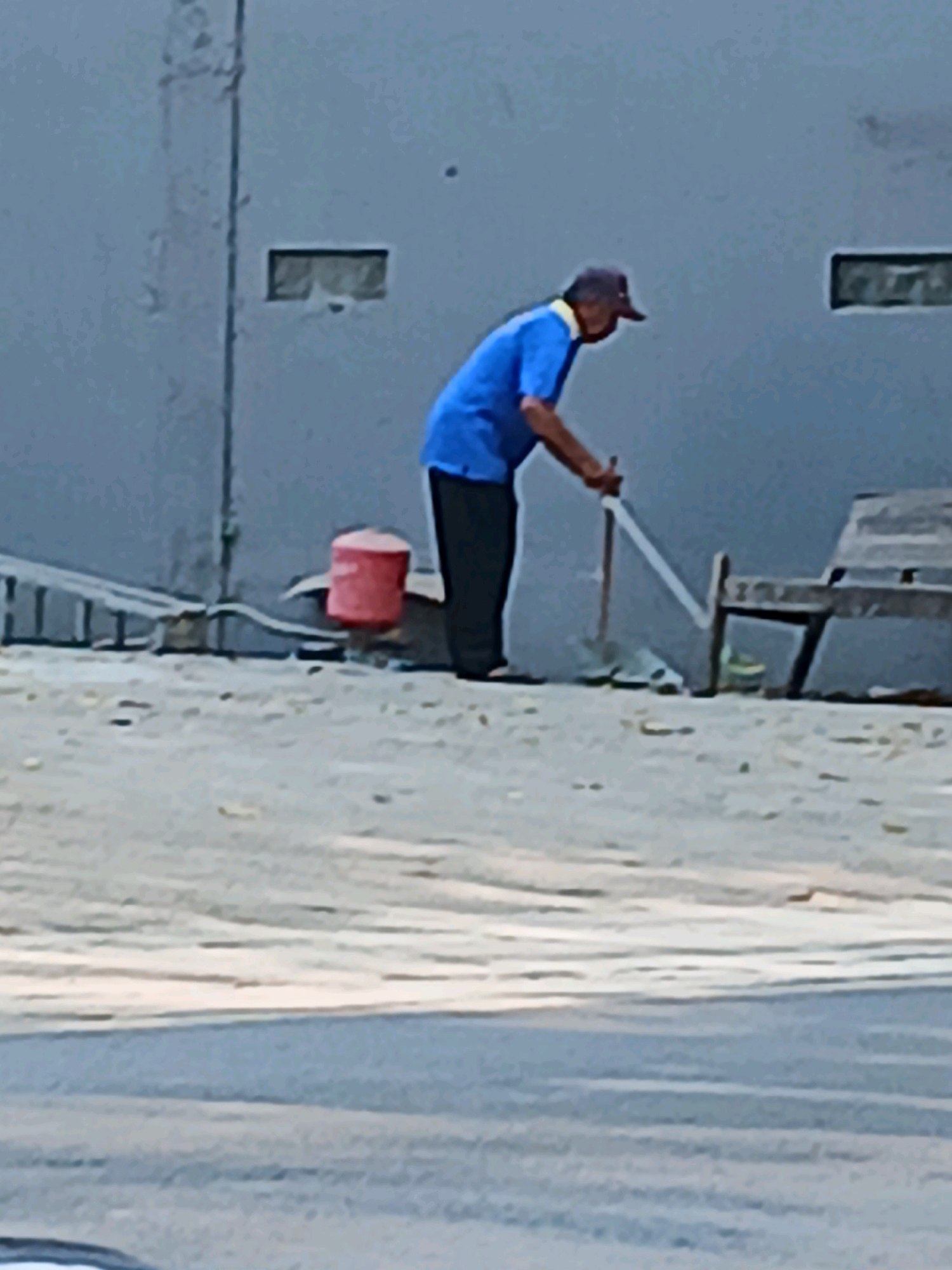 A man wearing a blue shirt and a dark cap is sweeping the ground with a broom. He is standing next to a red bucket and a wooden bench. The background is a gray wall with two rectangular openings. The scene appears to be outdoors, possibly in an urban environment in Kudus, Indonesia, given the architectural style of the wall and the presence of a bench often found in public areas. The ground is covered in sand and gravel.