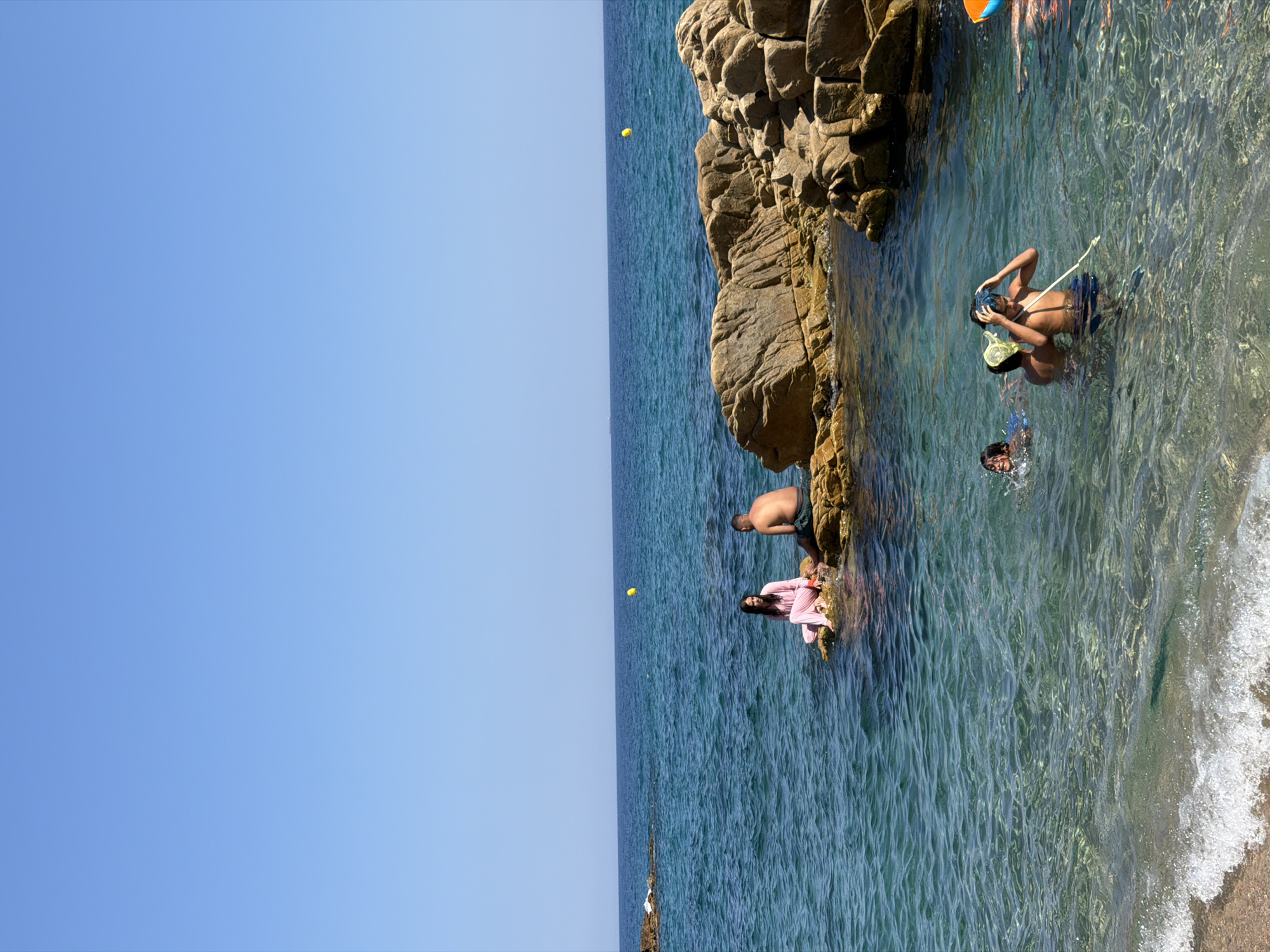 In the coastal city of Castell d'Aro, Platja d'Aro i s'Agaró, Spain, an idyllic scene unfolds on a sun-drenched day. The clear, turquoise waters of the Mediterranean Sea gently lap at a sandy shore, inviting a group of people to enjoy the refreshing embrace of the ocean.
Perched on large, weathered rocks near the shore, a man and a woman are captured in a moment of quiet contemplation. The woman, dressed in a flowing pink outfit, sits gracefully, her gaze directed towards the vast expanse of the sea. Beside her, a man, his torso bare, sits with his legs extended, also seemingly lost in thought as he observes the calming rhythm of the waves.
Further out in the water, a group of children engages in playful aquatic activities. Two boys are visible, one with a snorkeling mask and net, actively participating in a water-based pursuit. Another child is also in the water, their face reflecting a sense of pure enjoyment. The scene is further enhanced by the presence of a bright yellow buoy floating in the distance, a small marker in the grand canvas of the sea. The rugged, rocky coastline provides a dramatic backdrop to this tranquil seaside experience.