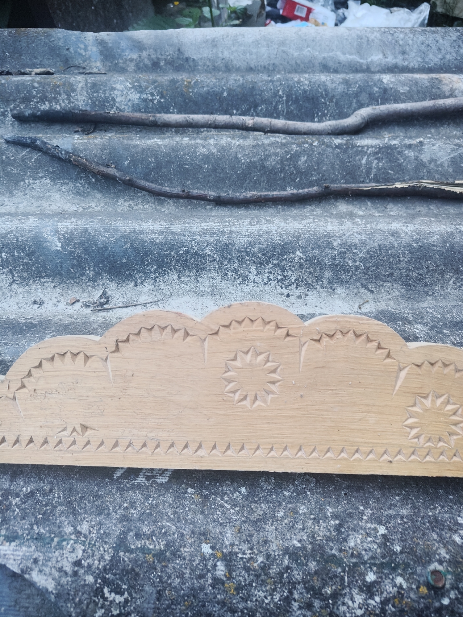 A carved wooden piece with a scalloped edge and star patterns rests on a gray, corrugated surface that appears to be roofing material. Two dry branches lie across the roofing material, one thicker and darker than the other. In the background, a pile of trash is visible, including plastic bags and a red and white box. The scene is outdoors, under natural light. The location is specified as Borova, Ukraine, suggesting this might be a detail observed in or around a household or workshop in that area.