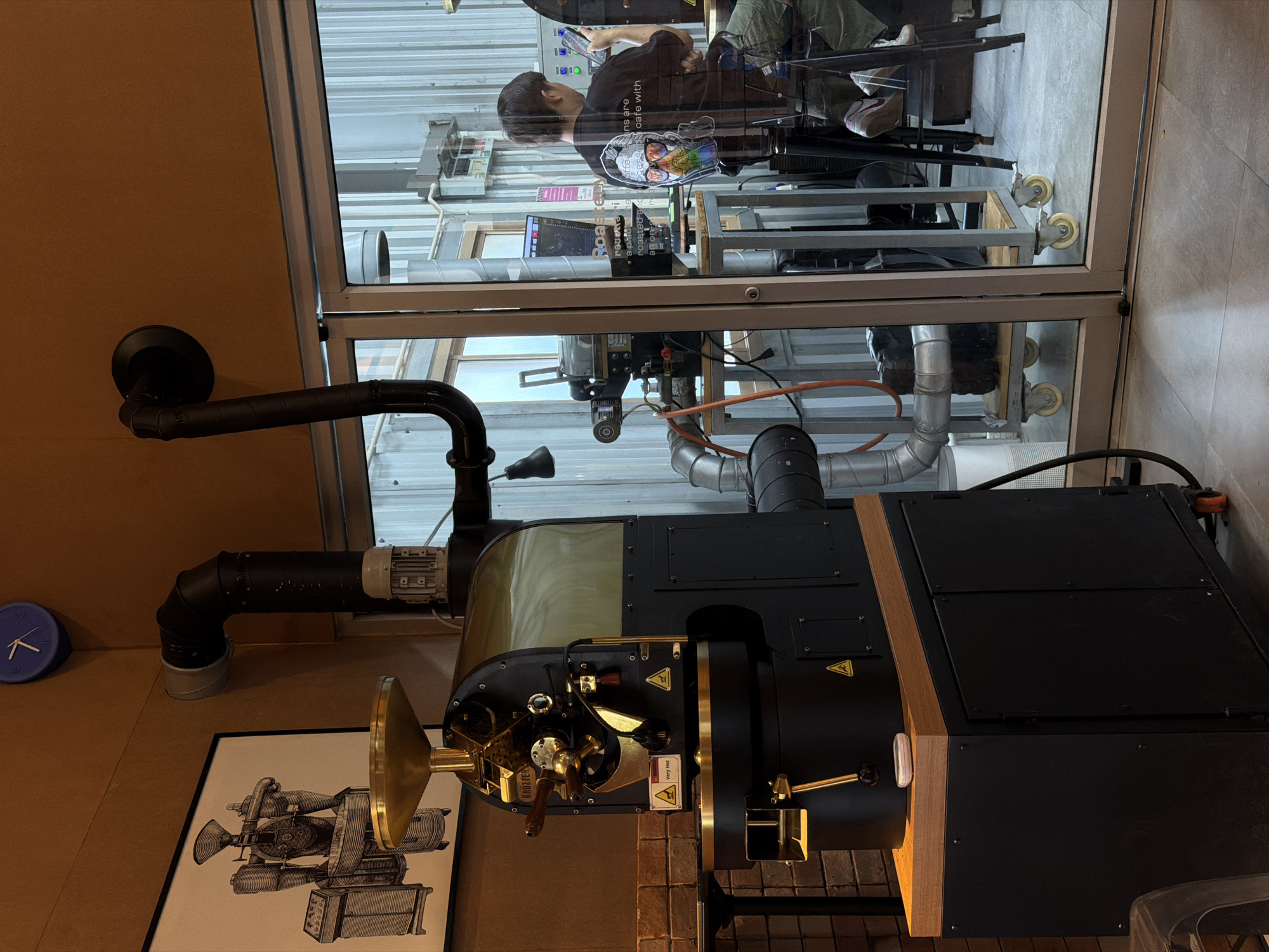 In the city of Chang Phueak, Thailand, a coffee roasting facility is depicted. A large, black and gold coffee roaster occupies the left side of the frame, with a gold funnel on top and an exhaust pipe system extending upwards and to the right. A framed illustration of a coffee roaster hangs on the wall behind it. To the right, through a glass partition, a person with dark hair is visible, wearing a black t-shirt with a colorful graphic and camouflage pants. They are seated and looking at a tablet, with their back mostly to the camera. In the background, various industrial elements and equipment are present, including metal structures, ventilation systems, and computers. The overall environment suggests a working coffee roastery.