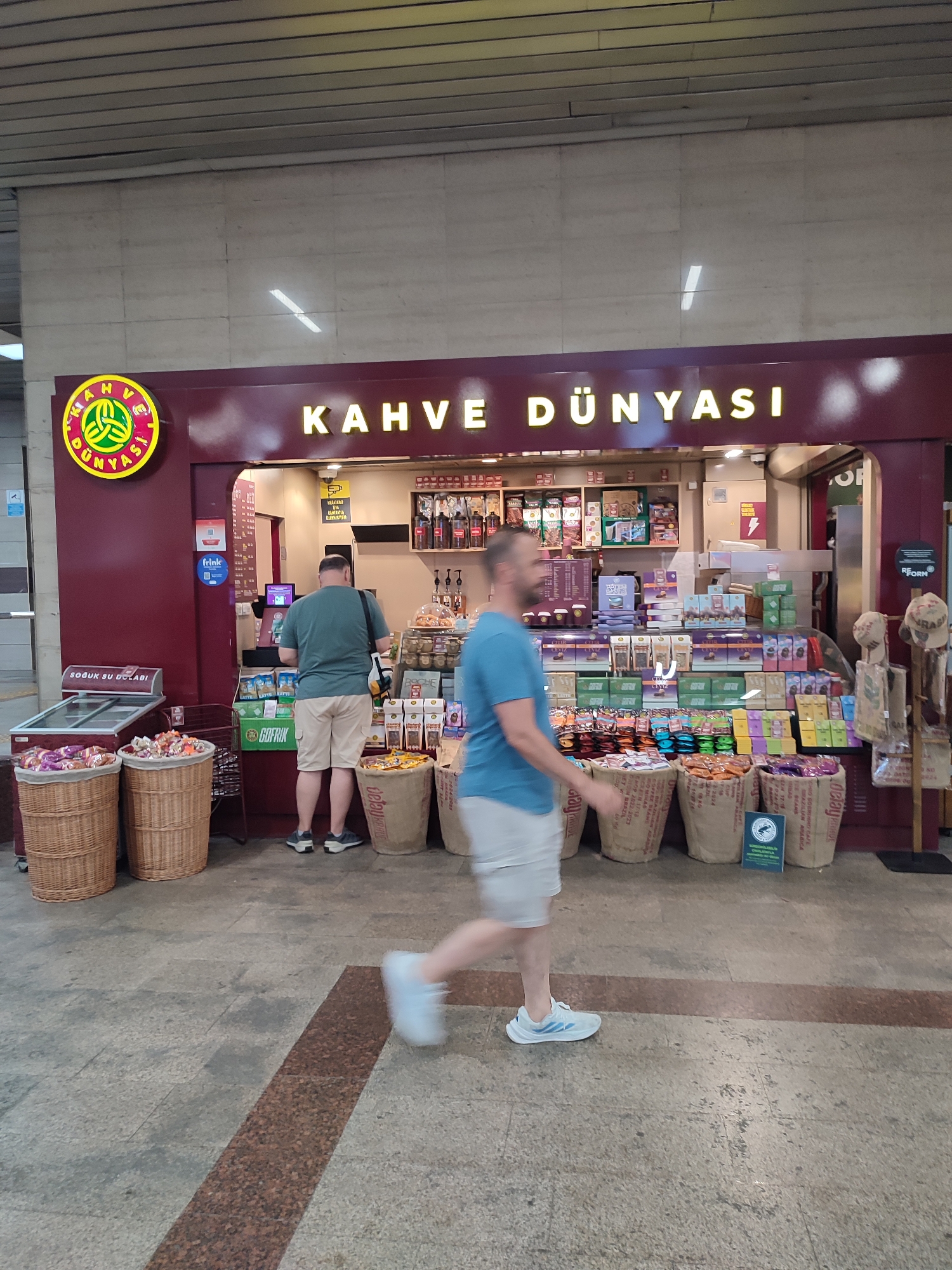The image depicts a street-level view of a "Kahve Dünyası" (Coffee World) establishment, likely a kiosk or a small shop, situated in a busy pedestrian area, possibly within the Beyoğlu district of Istanbul, Turkey, given the establishment's presence there. The foreground is dominated by a man in a blue t-shirt and white shorts, captured mid-stride and slightly blurred due to motion, walking away from the viewer and towards the right side of the frame. His light blue sneakers are clearly visible. To the left of this man, another man in a green t-shirt and beige shorts stands facing a counter, his back to the camera, appearing to interact with the vendor or browse products.
The "Kahve Dünyası" itself is characterized by a deep red facade with illuminated yellow lettering spelling out "KAHVE DÜNYASI". Above the entrance, a circular yellow sign with a green graphic is prominently displayed. The counter area is well-stocked with various coffee products, packaged snacks, and what appear to be beverages in bottles. Display shelves are filled with colorful boxes and bags of coffee and confectionery.
In front of the establishment, several large, woven baskets are placed on the ground. Two on the left are filled with colorful candies and wrapped goods. Three more on the right are filled with what appear to be coffee beans or similar bulk products, bagged in burlap sacks with visible branding. Adjacent to the baskets on the left, a glass-fronted refrigerated display case, labeled "SOĞUK SU DOLABI" (Cold Water Cabinet), contains items.
The background shows the interior of the shop with more shelves and a security camera visible. The flooring is a textured grey stone with a reddish-brown speckled strip running diagonally across the frame in the foreground. The overall impression is that of a bustling commercial environment, typical of a popular coffee chain in a Turkish city.
