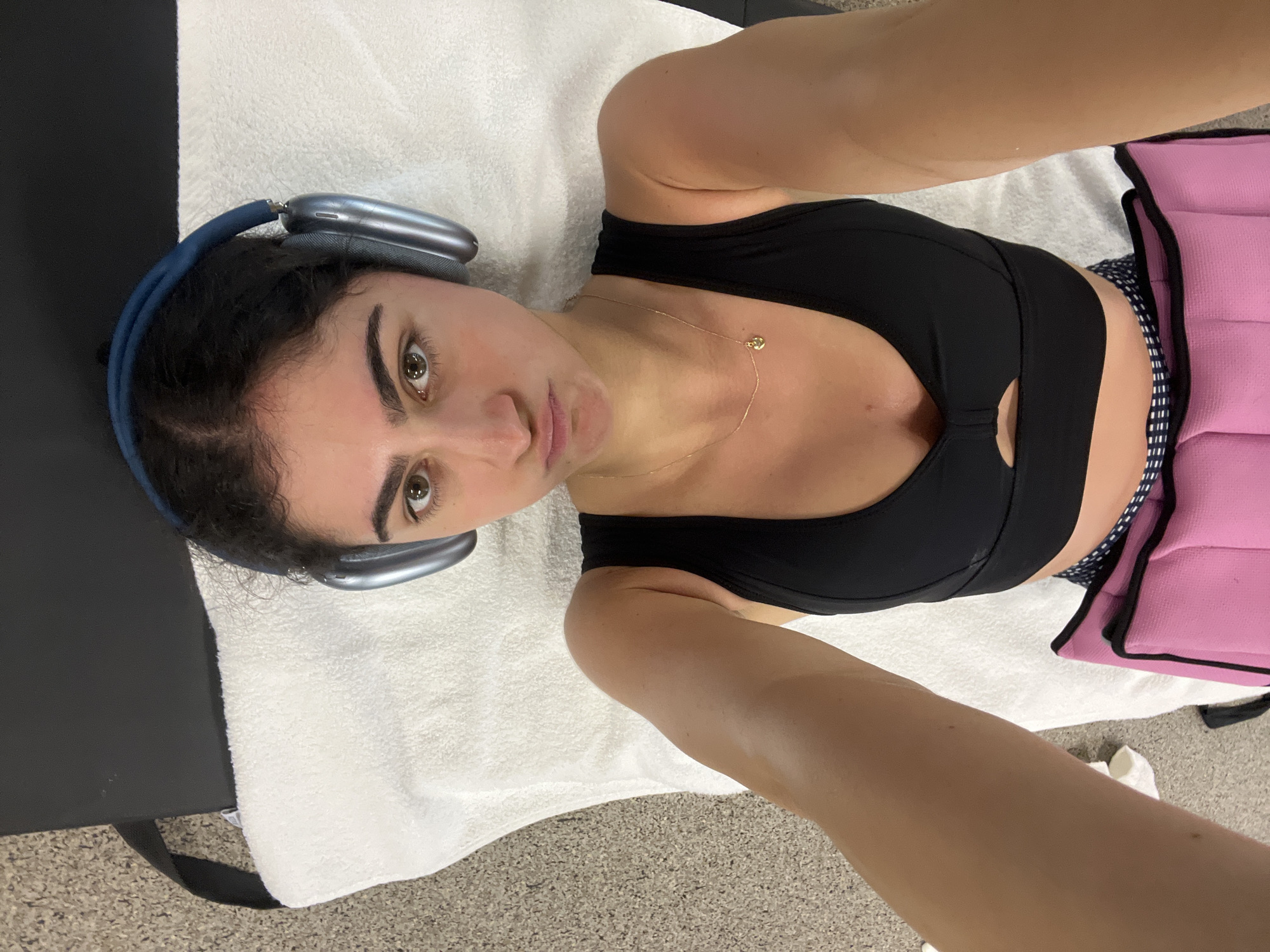 A woman is lying on a white towel on what appears to be an examination table. She is wearing headphones and a black sports bra. A pink weight bag is visible near her waist. The scene is indoors and the location might be in the city of Miami Beach, United States of America.