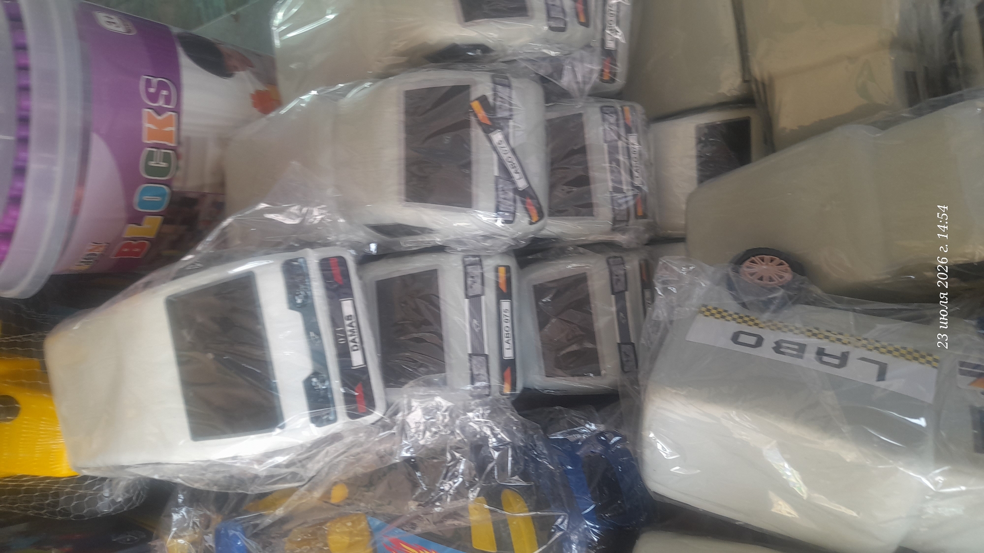 This is a close-up, eye-level, indoor photograph of several toy vehicles, likely a fleet of small, white vans, neatly arranged and individually wrapped in clear plastic. The toys are the primary focus, dominating the frame. Some of the toy vehicles have labels on their front grills. One label reads "0/1 DAMAS," and another reads "LABO 075." The toys are stacked or placed side-by-side, suggesting they are either for sale or storage. In the background, partially visible, is a large, purple and white plastic tub with the word "BLOCKS" printed on it in colorful letters, suggesting it might contain toy building blocks. The lighting is somewhat dim, and the overall impression is that of a commercial setting, possibly a shop or warehouse in Shurchi, Uzbekistan. The timestamp "23 июля 2026 г. 14:54" is superimposed on the bottom right of the image, indicating the date and time the photograph was taken.
