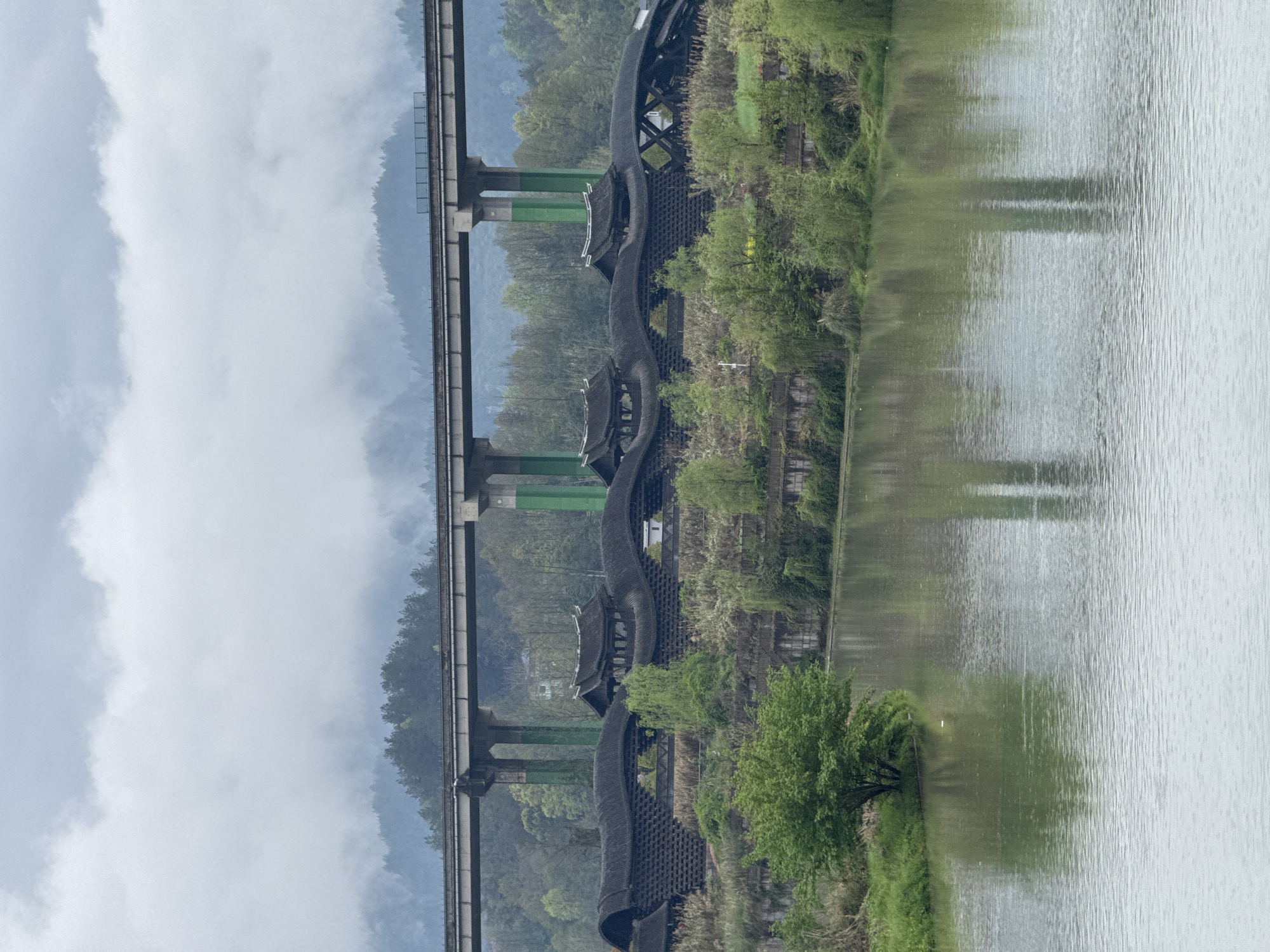 The image depicts a scene in Zhuoshui, China, featuring a distinctive covered bridge known as the Shuanglong Bridge, situated adjacent to a modern elevated railway bridge. The bridges span a body of water, presumably a river or lake. The covered bridge, of traditional Chinese design, showcases a serpentine structure with ornate roof elements. Lush greenery is present along the riverbank, reflected in the water. The railway bridge is supported by green pillars. The sky is overcast.