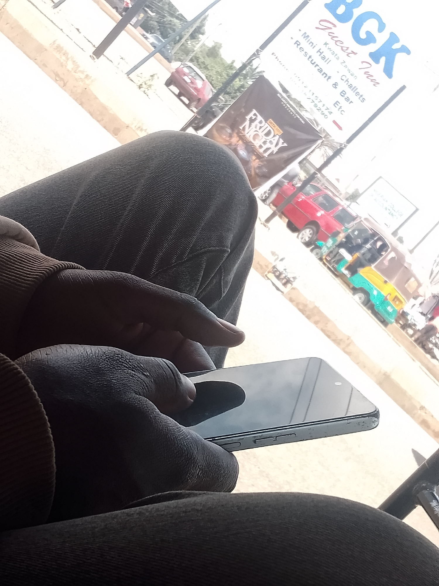 A person is seated, holding a smartphone. The scene is outside, with a "BGK Guest Inn" sign visible, along with other signs and advertisements. Several vehicles are visible in the background including cars and a tricycle. The guest inn is located in Kwata Zawan along Vom Road.