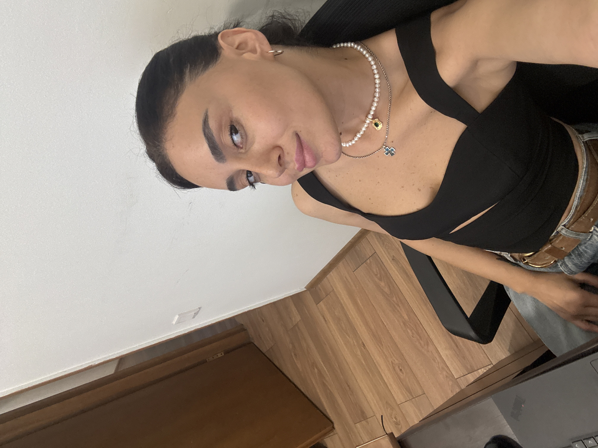 A young woman with dark hair pulled back is captured in a high-angle, indoor selfie. She is seated in a black office chair at a desk with a laptop. The woman is wearing a black, cut-out crop top, blue jeans, and a brown leather belt. She accessorizes with a pearl necklace featuring two pendants and small hoop earrings. The background shows a wooden floor and a partially open wooden door leading into another room. The wall behind her is painted white, with a light switch visible on the left. The lighting is natural, coming from the left side of the frame.