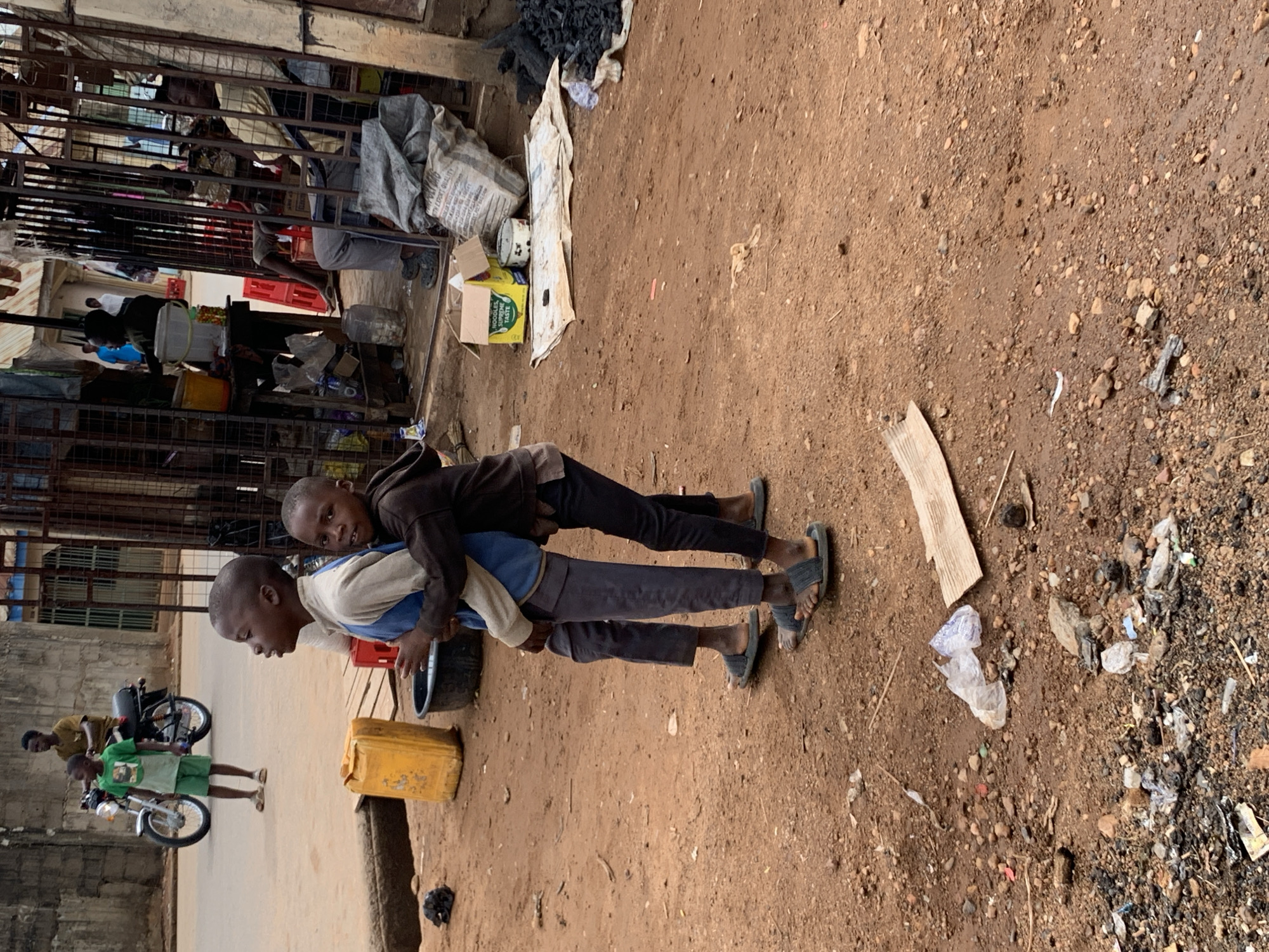 Two boys are giving each other piggyback rides in an outdoor area of Akwanga, Nigeria. One boy wears a brown hoodie and black pants, while the other has a light-colored shirt and dark pants. Both are wearing sandals. A yellow jerrycan is on the ground nearby. In the background, a motorcycle is parked on the side of the road, and a third person is standing beside the motorcycle. Behind the children is a building or structure with various items stacked or stored inside. The ground is dirt with scattered debris.