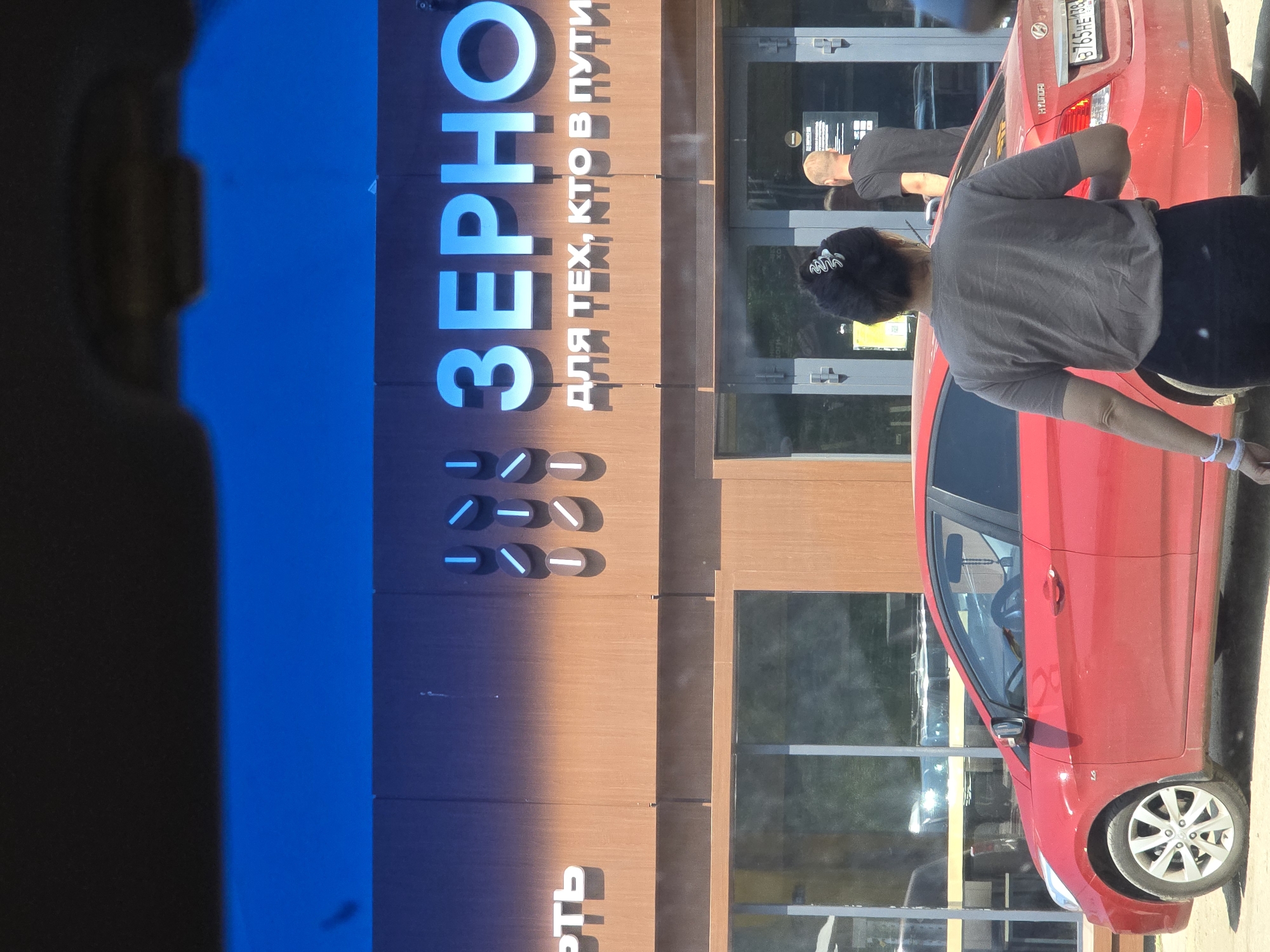 In Turka, Russia, a red Hyundai Accent is parked in front of a building with a brown facade. Above the building's entrance, large blue letters spell out "ЗЕРНО" (Zerno), which translates to "grain." Below this, smaller white text reads "ДЛЯ ТЕХ, КТО В ПУТИ" (Dlya tekh, kto v puti), meaning "For those on the go." To the left of "ЗЕРНО," a stylized logo composed of vertical and diagonal lines within circles is visible. A woman wearing a grey t-shirt and black pants, with her hair in a bun, stands near the car. In the background, a man wearing a black t-shirt stands at the entrance of the building. The sky above is a bright, clear blue.