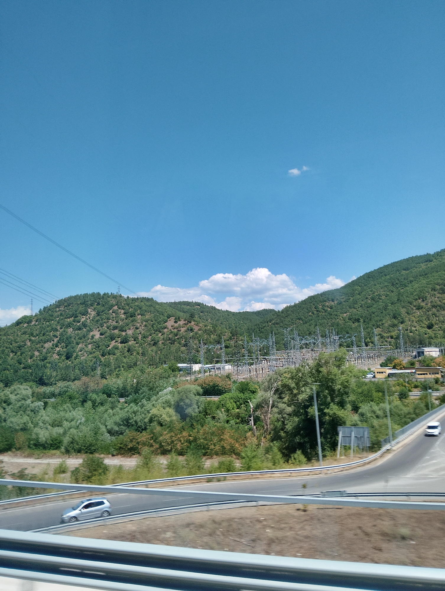 The image shows a view from a moving vehicle on a highway in Blagoevgrad, Bulgaria. In the foreground, a silver car is visible on the road. The highway curves to the right, with guardrails on either side. In the middle ground, there is a vast power substation with numerous metal towers and electrical equipment, nestled at the base of a lush green, tree-covered mountain. The mountain slopes upwards, disappearing into the bright blue sky, which has a few wispy white clouds. Power lines stretch across the sky and ascend the mountainside, connecting to tall pylons. The overall scene depicts a blend of industrial infrastructure and natural landscape.