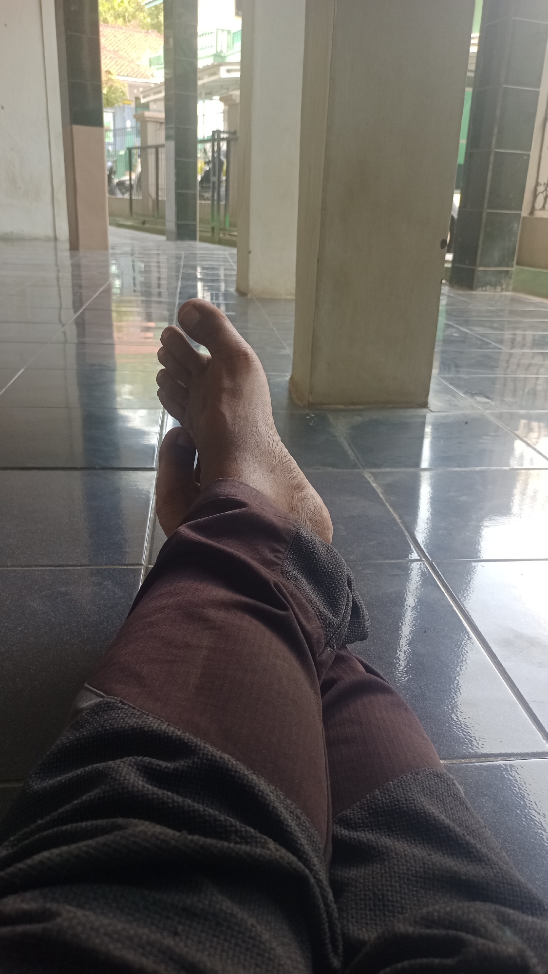 A person's feet and legs are visible, lying on a reflective tiled floor. The person is wearing dark brown pants with a textured dark grey trim at the ankles. The floor tiles are large and dark, reflecting the light from overhead and from the environment visible in the background. Behind the person, several large, light-colored pillars are present, suggesting an indoor or covered outdoor space. In the distance, through the gaps between the pillars, a street scene is visible, with buildings, a fence, and some vehicles. The overall impression is a casual resting pose in a public or semi-public area.