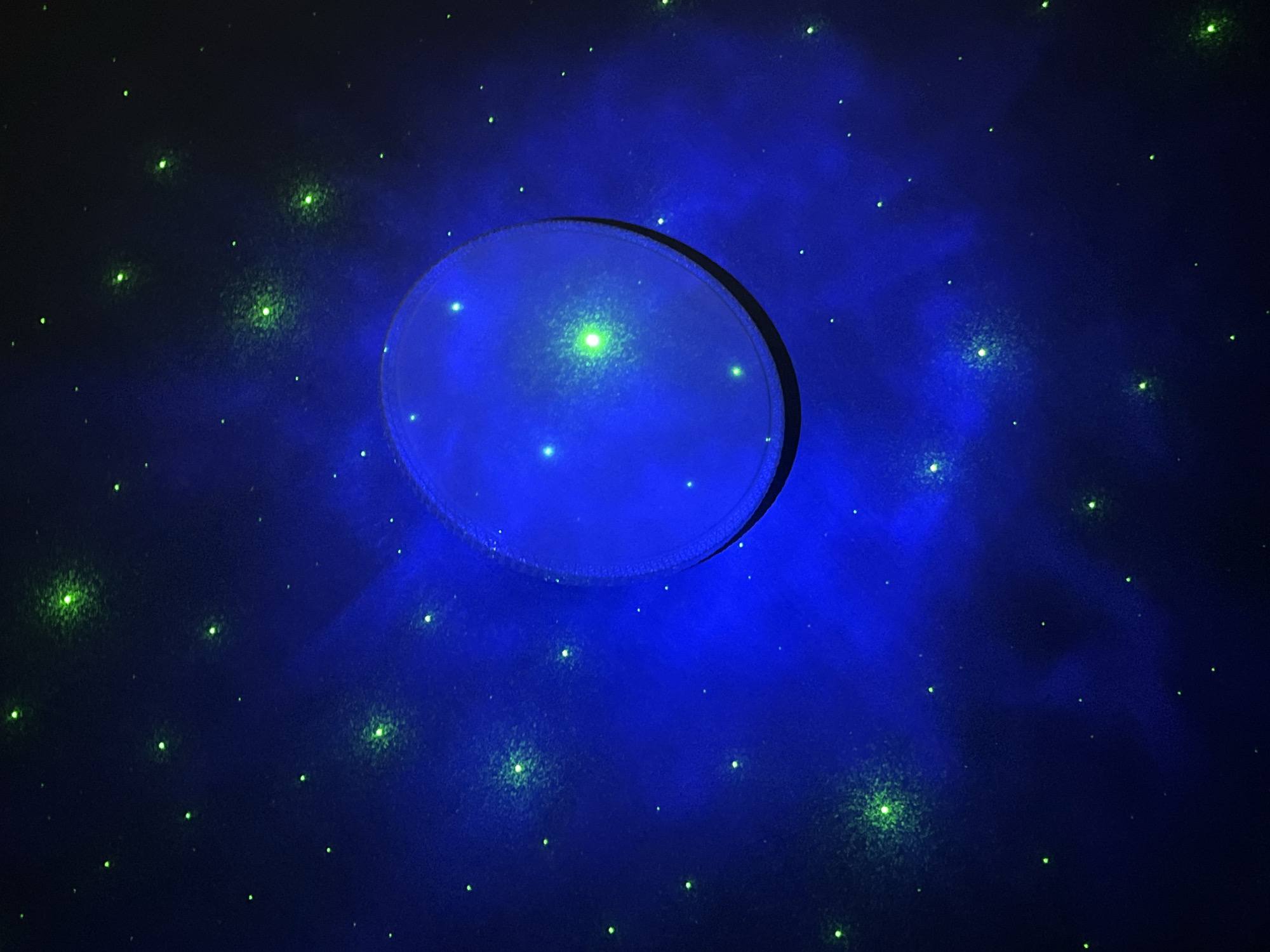 A circular object is positioned in the center of the image, its surface a vibrant blue with lighter blue points distributed across it. A dark border encircles the circle. The background is a deep blue, resembling a night sky, with numerous small, green lights scattered throughout, mimicking stars. The overall scene evokes a celestial or astronomical theme. The image does not contain information that would support the location in the city of Nizhny Tagil, Russia.