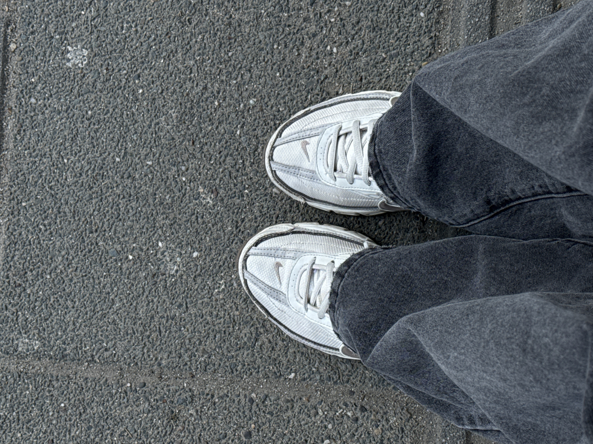 A top-down shot shows a person standing on a paved asphalt surface. The individual wears grey wash denim jeans that obscure most of their legs. The feet are clad in white Nike sneakers with matching laces. The asphalt is grey and textured. The location is not discernible from the image.