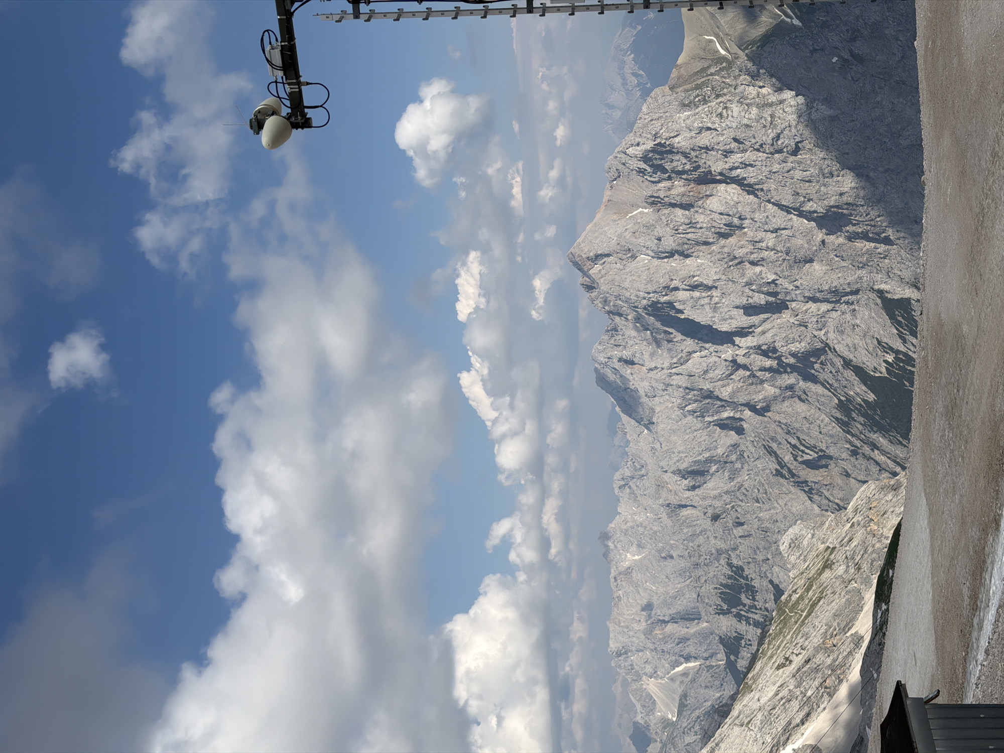 This image depicts a mountainous landscape, likely taken from a high vantage point. The scene features rugged, grey mountain peaks with sharp inclines and visible rock formations. Patches of snow or ice can be seen on some of the higher slopes, suggesting a cool, alpine environment. The sky is blue with scattered white cumulus clouds, indicating fair weather.
In the foreground, a section of a metal structure is visible at the top of the frame, possibly part of a cable car system or observation deck. Attached to this structure are what appear to be antennae and possibly some lighting fixtures. To the right of the mountains, a textured, light-colored surface dominates the edge of the frame, which could be a wall or the side of a building.
The orientation of the image is unusual, with the mountain range appearing to run vertically. This suggests the photo may have been taken with the camera rotated. The lighting indicates it is daytime, likely mid-morning or afternoon, given the bright sky and the shadows cast on the mountainside. The overall mood is one of awe at the natural grandeur of the mountains, with a hint of human presence indicated by the technological equipment. There is no visible text in the image. The location context of Garmisch-Partenkirchen, Germany, aligns with the visual cues of a high-altitude alpine setting.