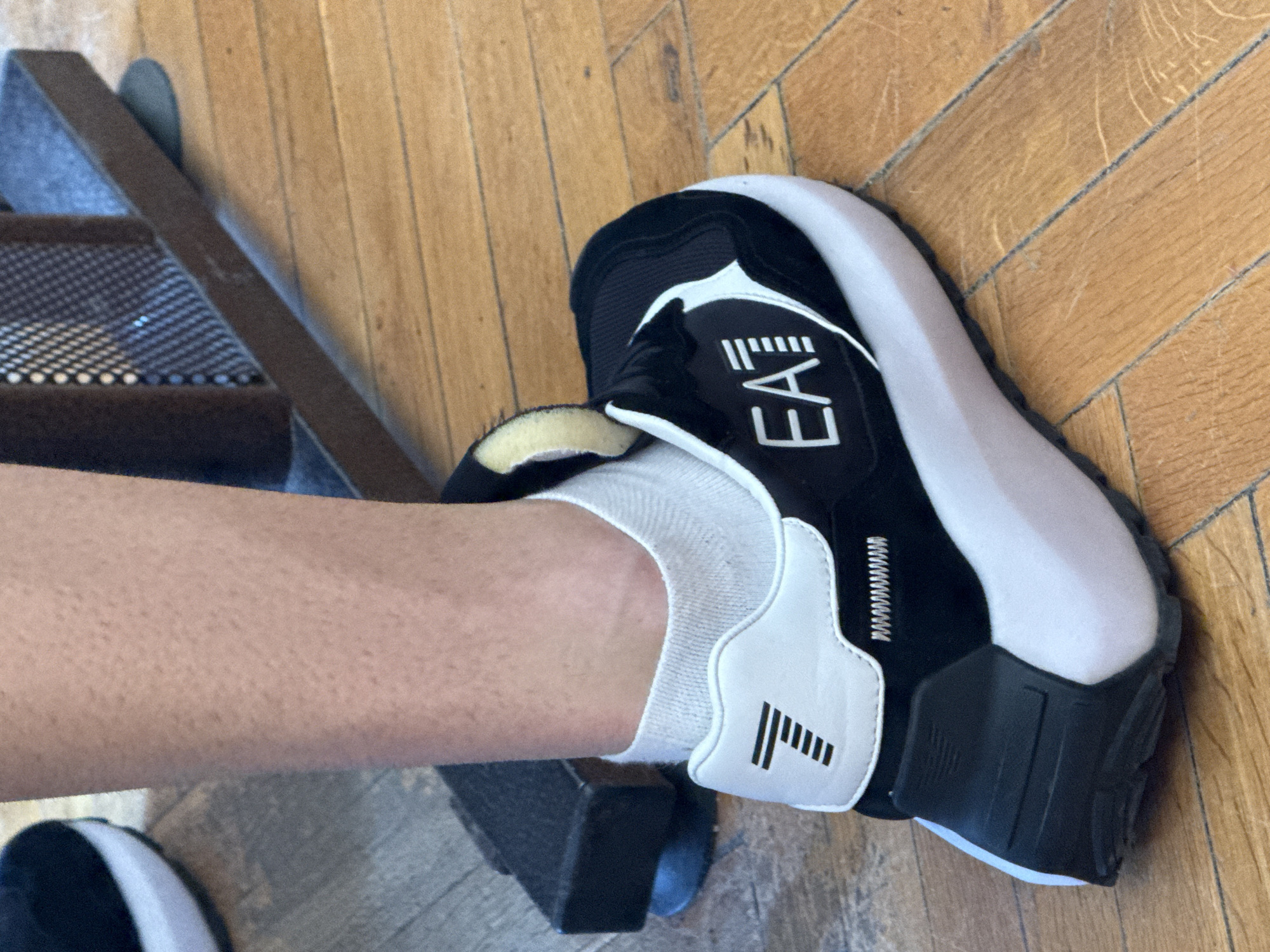 A close-up photograph shows a person's leg and foot, wearing a white sock and a black and white EA7 Emporio Armani sneaker, standing on a wooden floor next to a black metal step. The location cannot be determined as Elhovo, Bulgaria based on visual evidence.